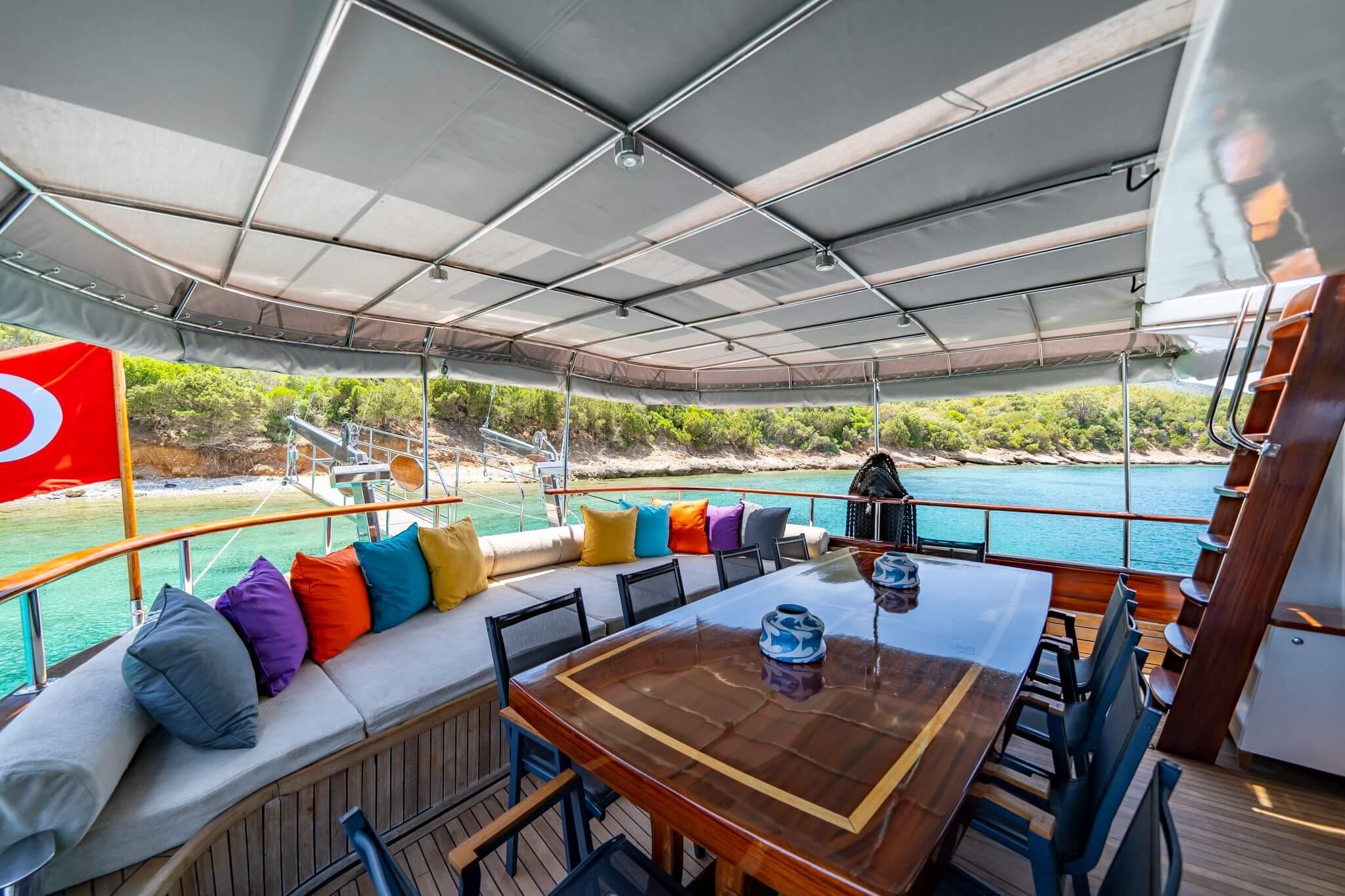 Gulet aft deck with large wooden dining table, colourful cushioned curved sofa, and retractable awning canopy overlooking turquoise cove