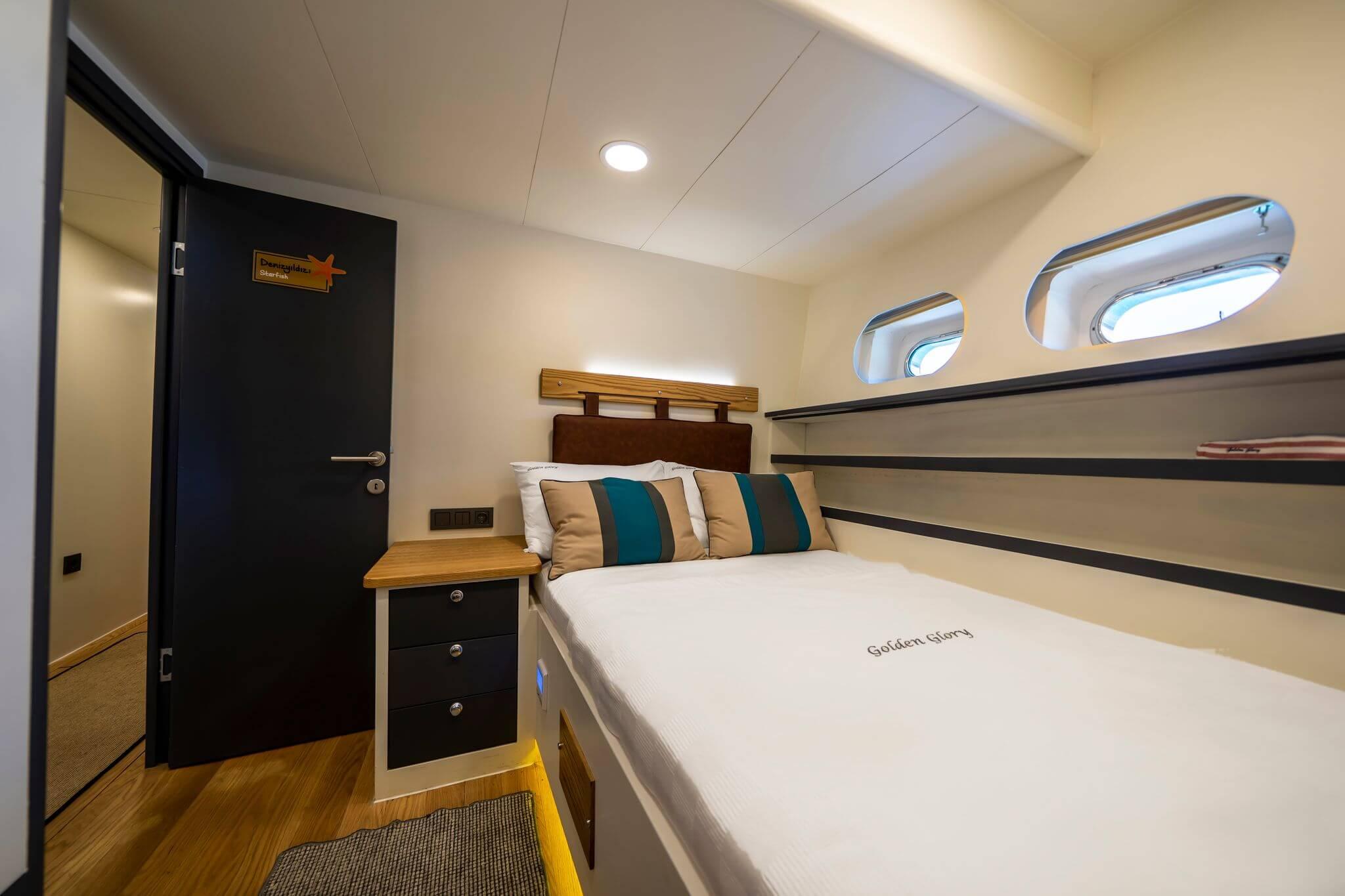 Double cabin on gulet Golden Glory with white bedding, teal striped pillows, wood headboard, and two oval portholes