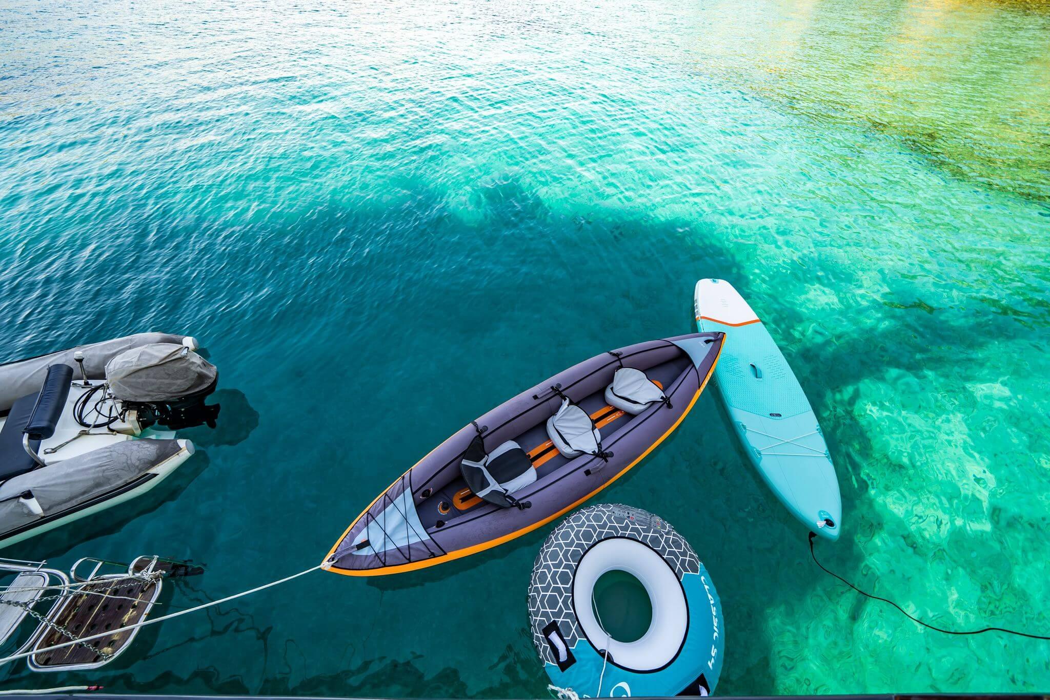 Water toys including inflatable two-seat kayak, SUP paddleboard, and inflatable ring float alongside yacht tender in crystal-clear turquoise water