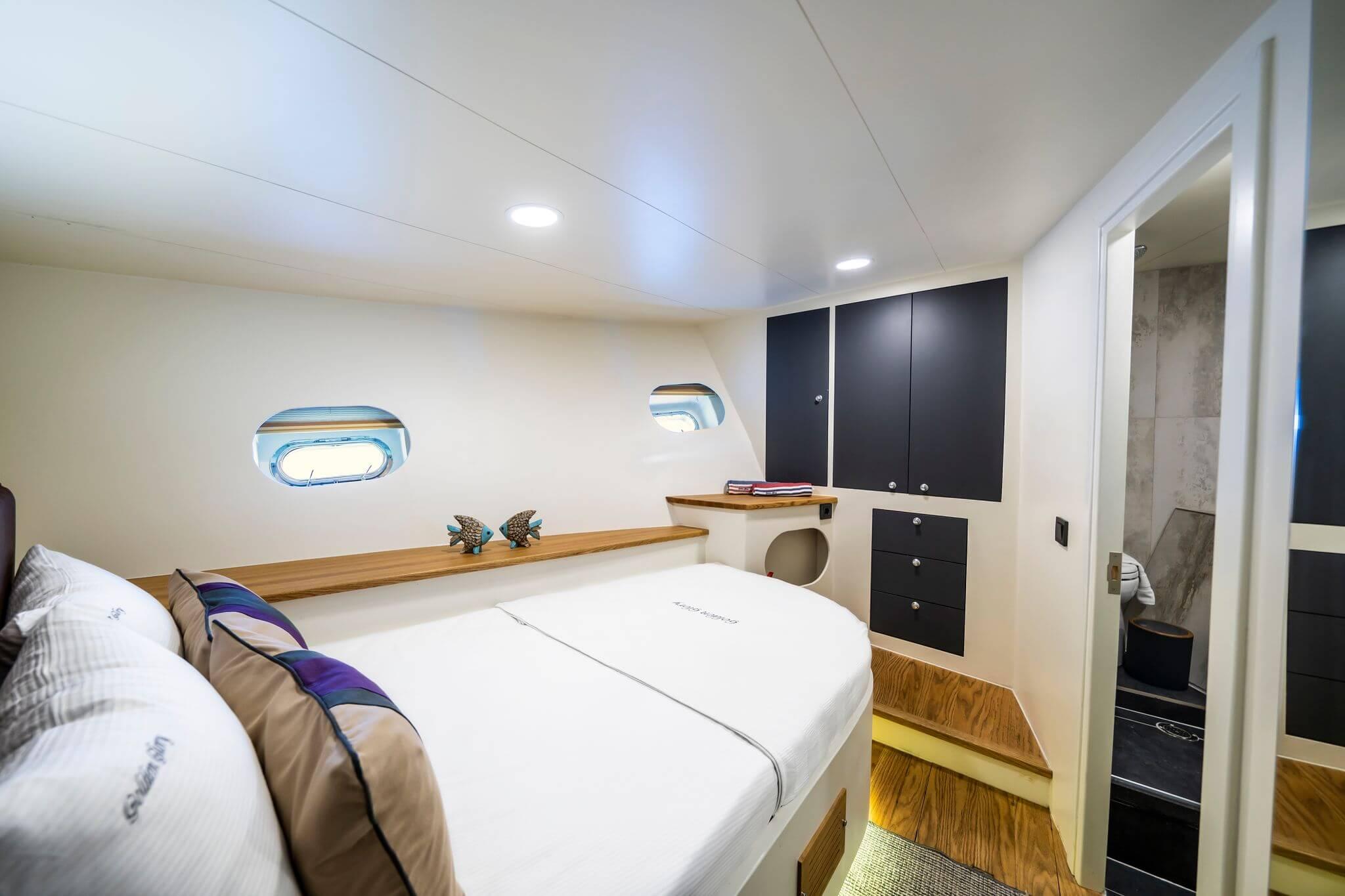 Yacht double cabin with white bedding, two oval portholes, teak shelf, and dark navy storage cabinets with en-suite visible