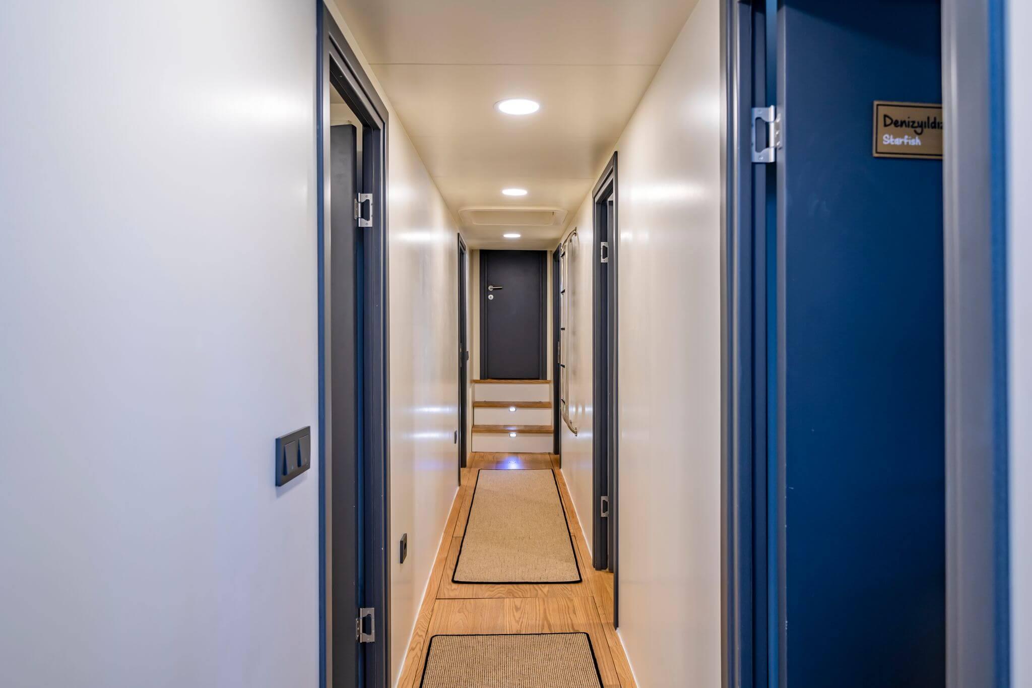 Below-deck passageway with multiple named cabin doors, wood flooring, recessed lighting, and staircase at end