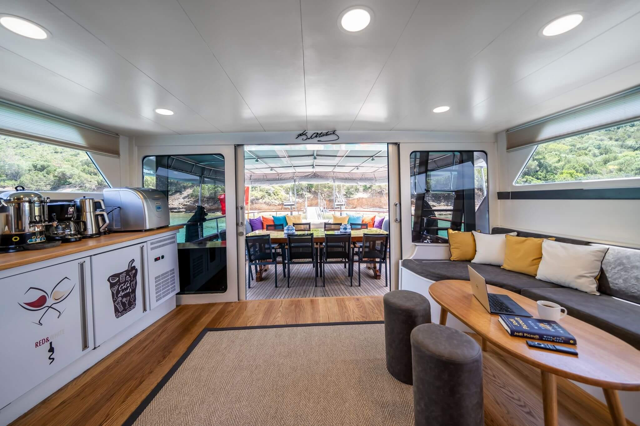 Gulet saloon with espresso bar, grey sofa, and open aft deck dining table with colourful cushions