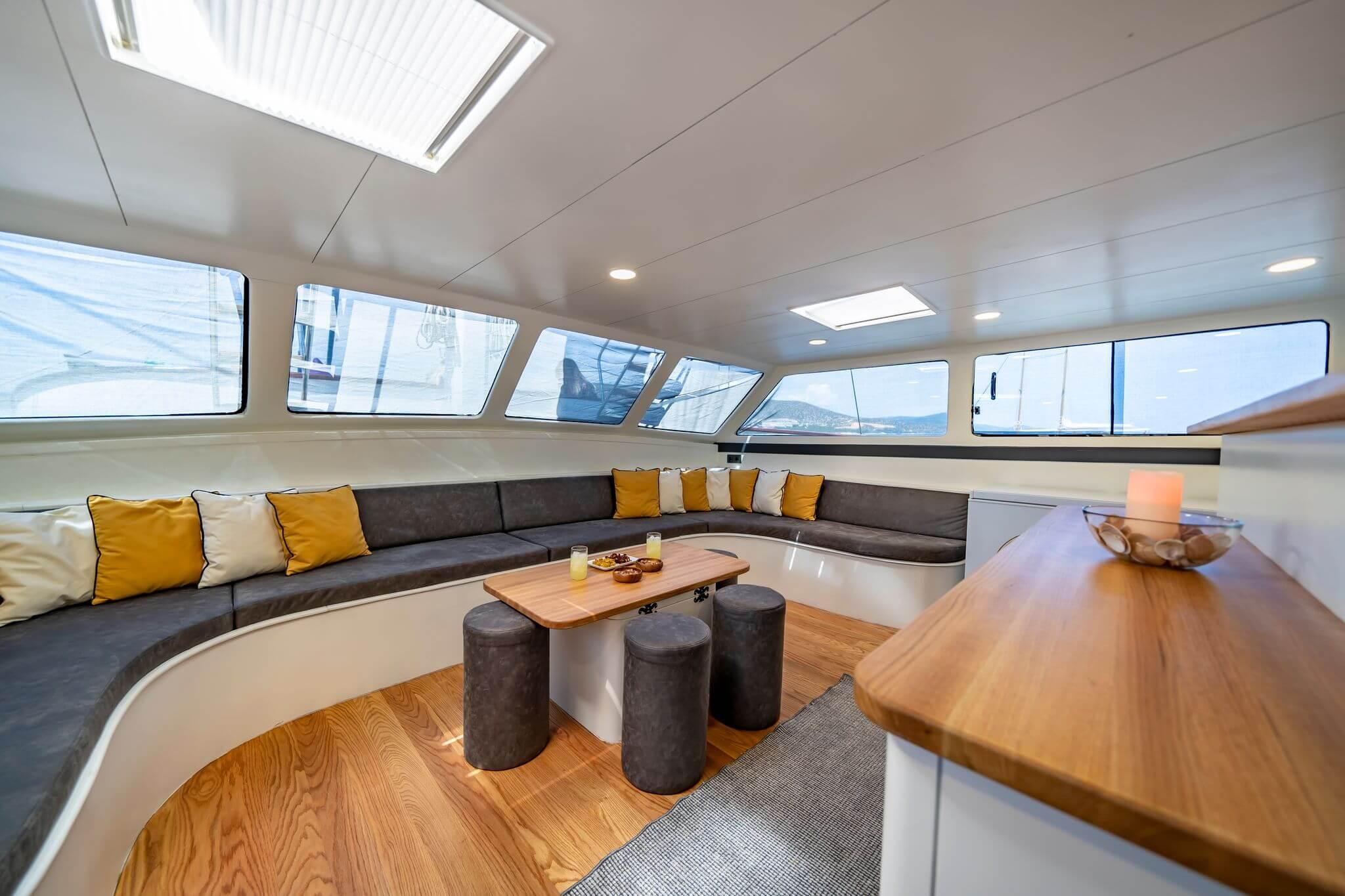 Gulet interior saloon with curved dark grey sofa, yellow cushions, low teak coffee table with drinks and snacks, and panoramic windows