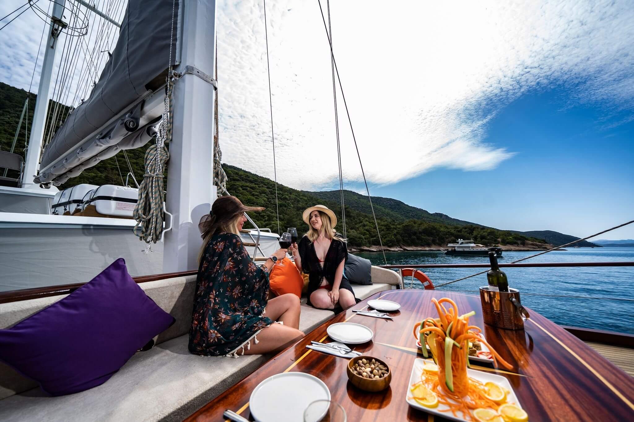 Two women clinking wine glasses at a polished wooden deck table with fresh fruit platters and snacks, anchored in a calm bay