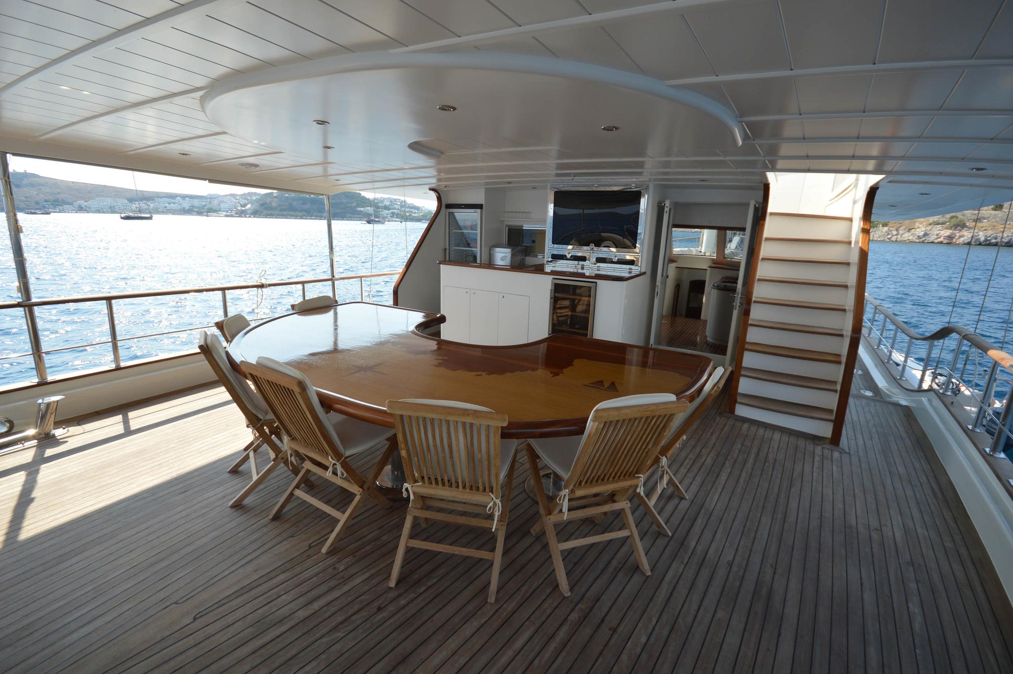 yacht aft deck dining area with round wooden table, teak chairs and galley kitchen