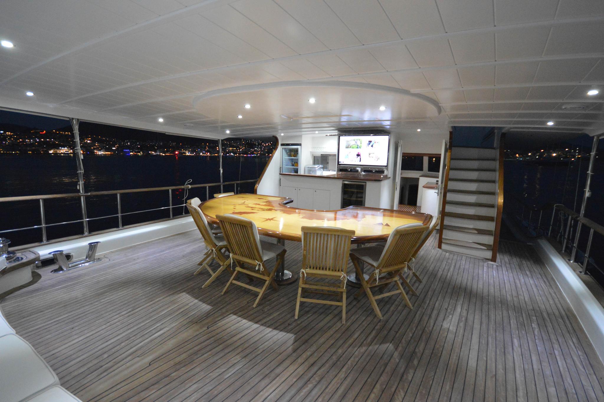 yacht aft deck dining area with wooden table, teak chairs and galley kitchen at night