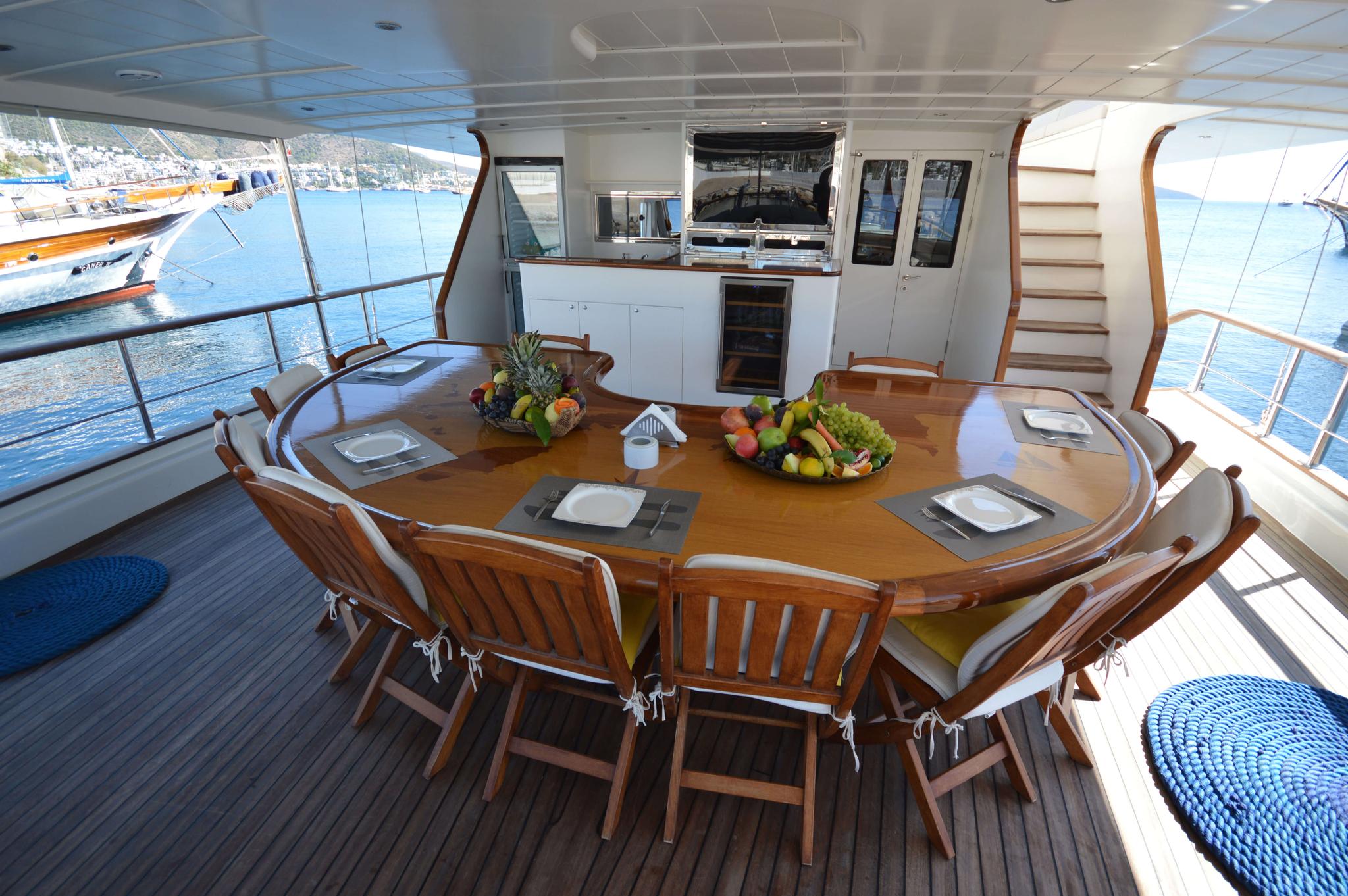 Yacht aft deck dining area with oval teak table, wooden chairs, and galley kitchen behind