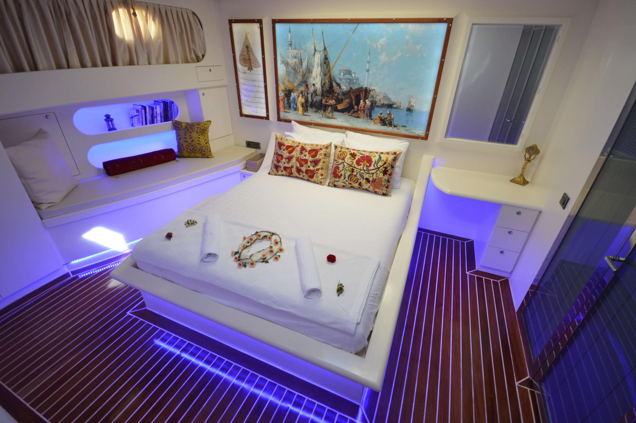yacht cabin interior with white double bed, blue LED lighting, maritime artwork and teak flooring