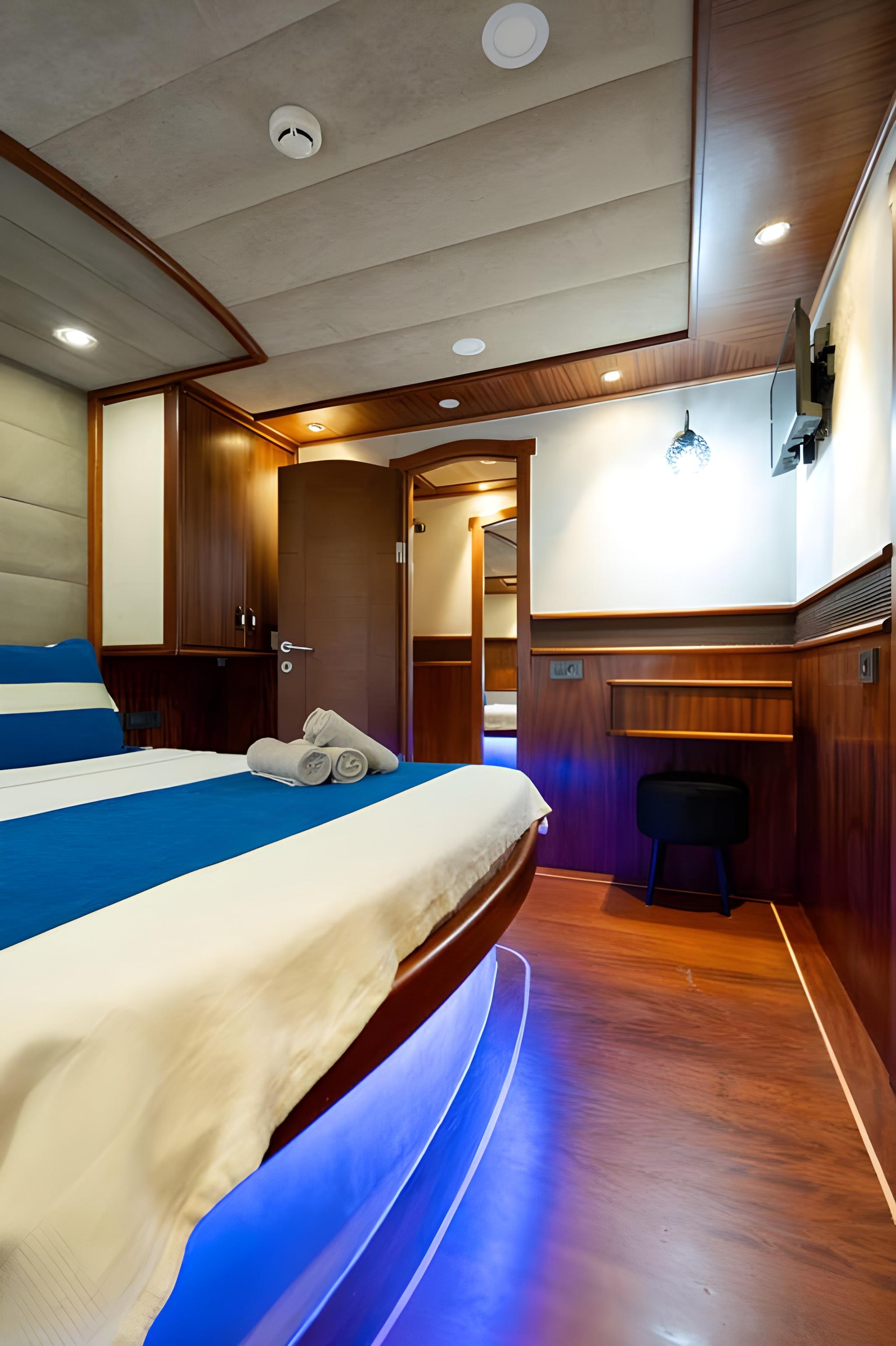 yacht guest cabin with double bed, white linens, dark wood paneling and blue accent lighting