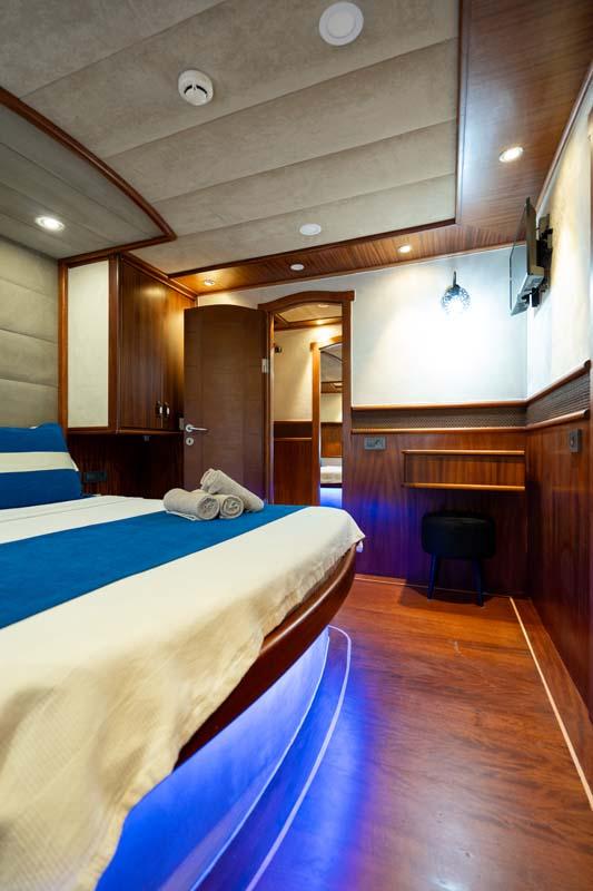 yacht guest cabin with double bed, white linens, dark wood paneling and blue accent lighting
