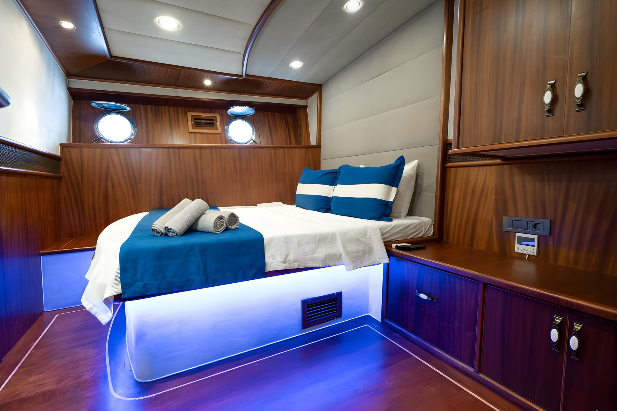 yacht cabin interior with double bed, blue accent lighting, portholes, and wooden cabinetry