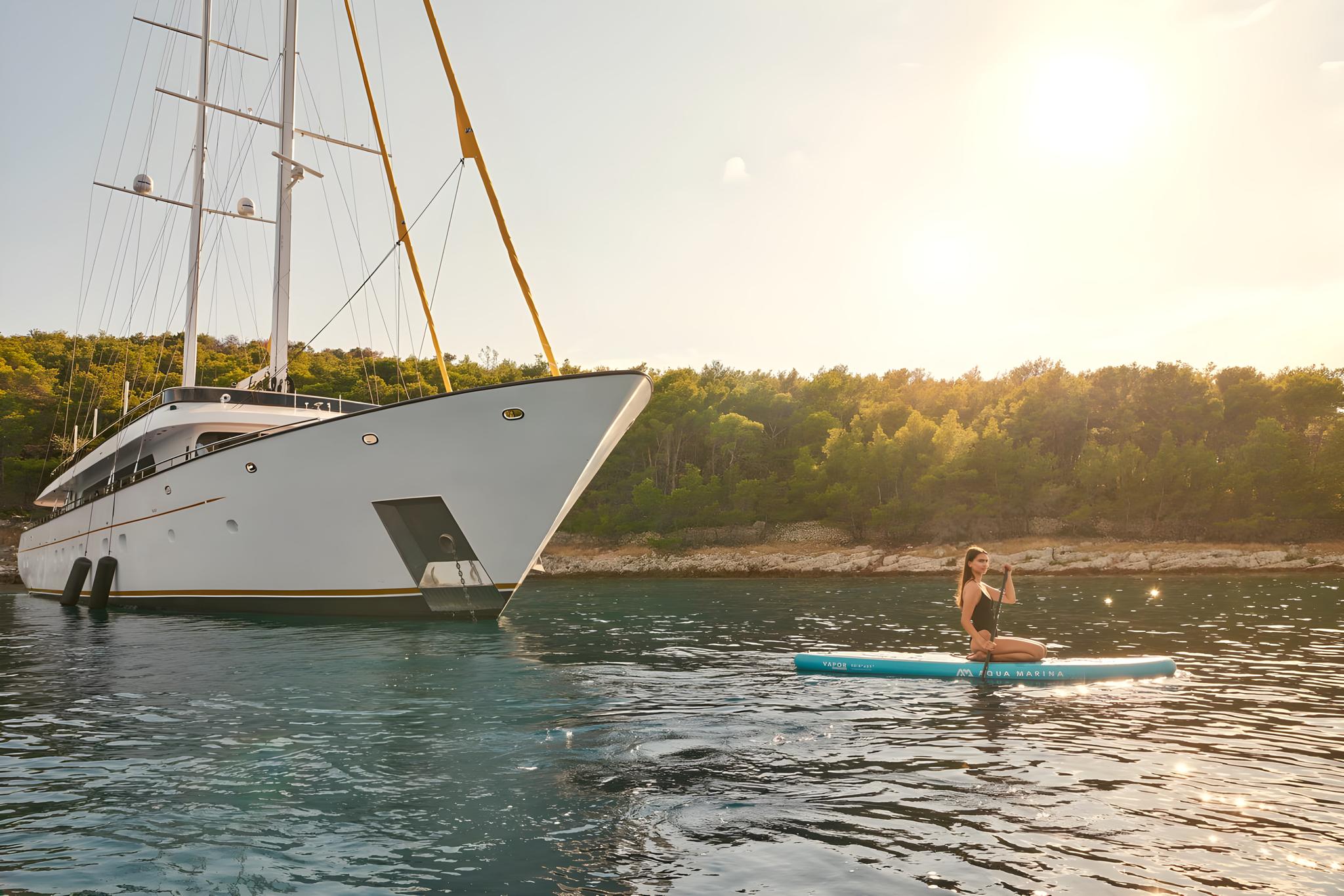 White luxury sailing yacht anchored in turquoise waters with woman on paddleboard