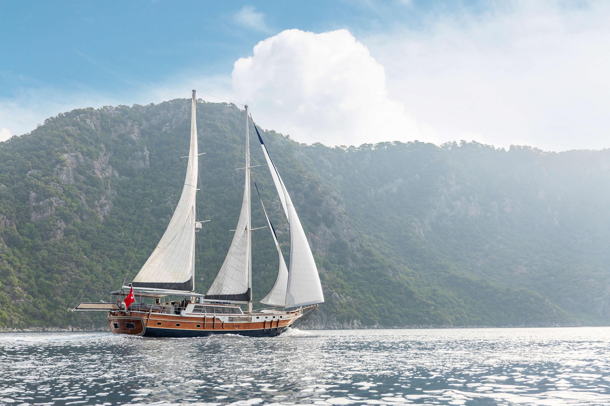 Traditional wooden gulet under sail with white sails against forested mountainous coastline