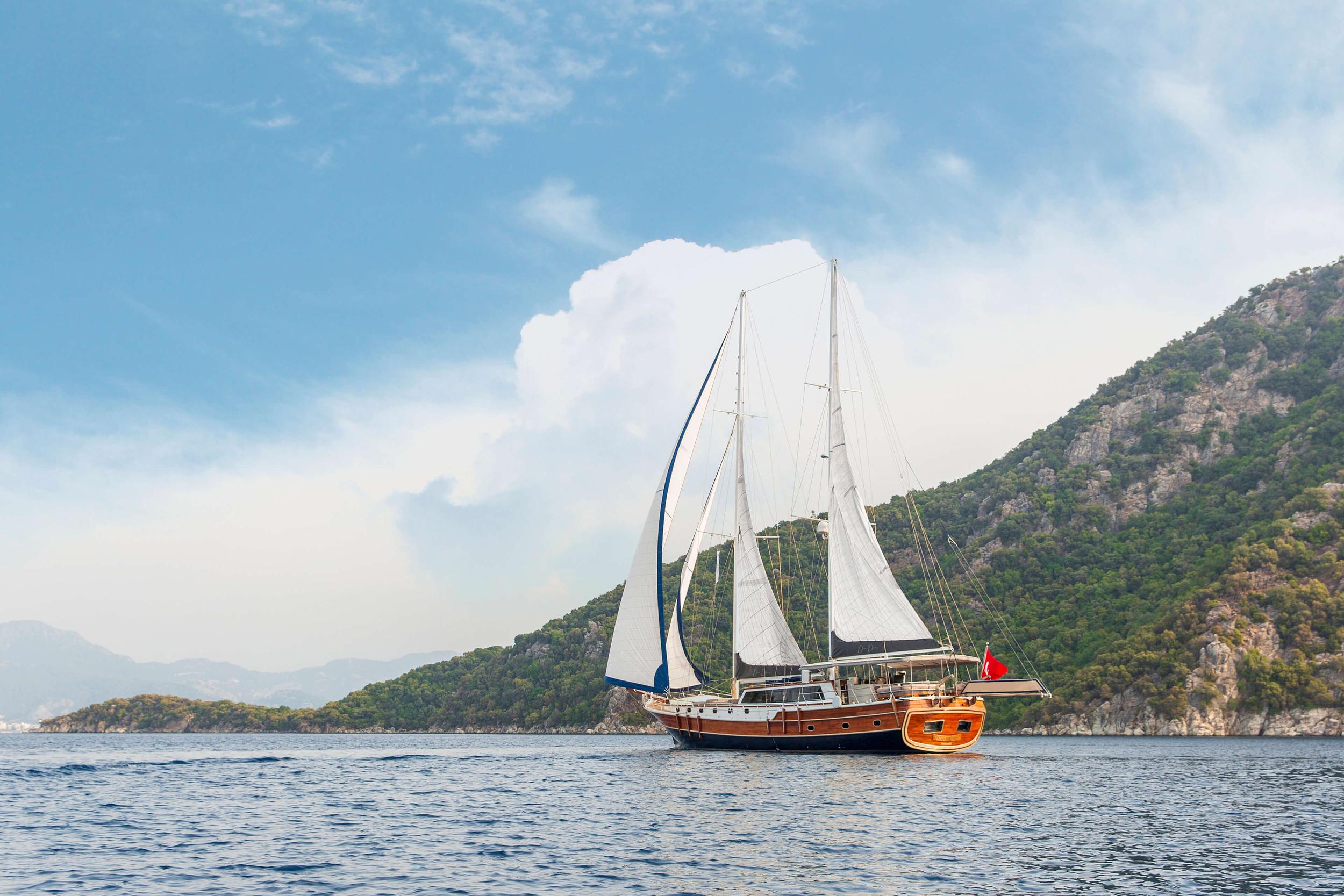 Traditional wooden gulet yacht sailing with white sails against mountainous Mediterranean coastline