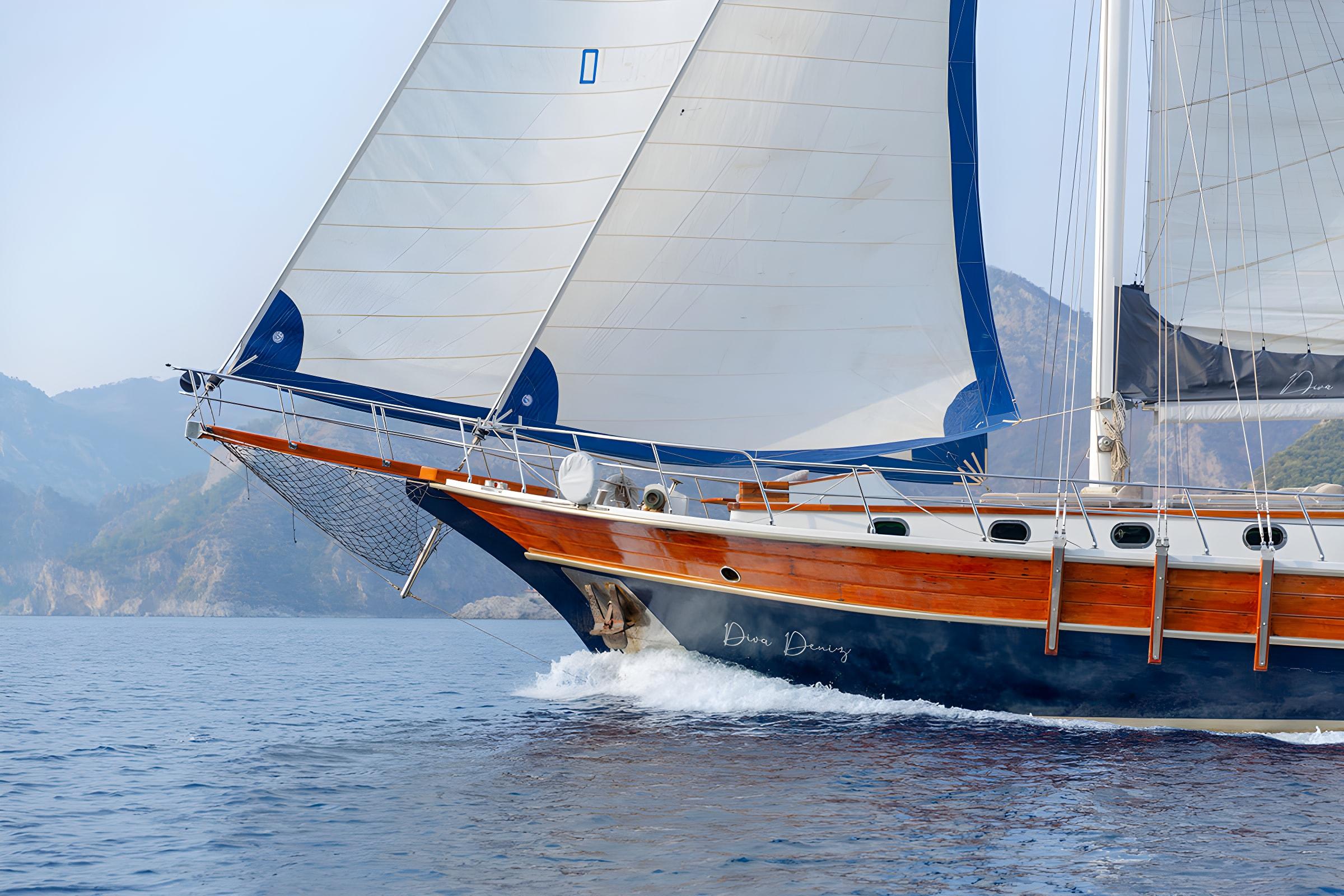 wooden gulet Diva Deniz sailing with blue and white sails raised against mountainous coastline