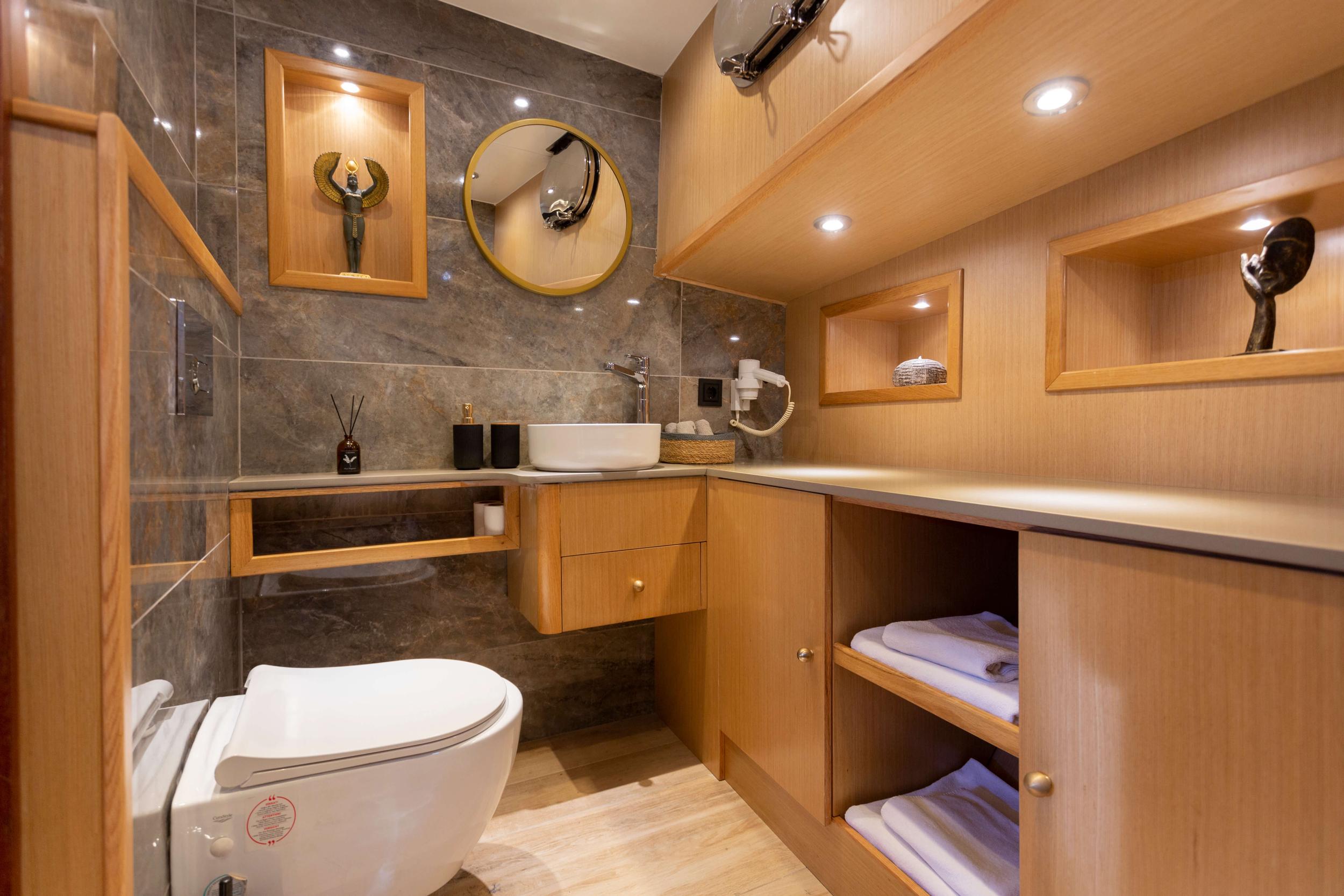 yacht bathroom with white vessel sink, gray marble tiles, wooden cabinetry and towel storage