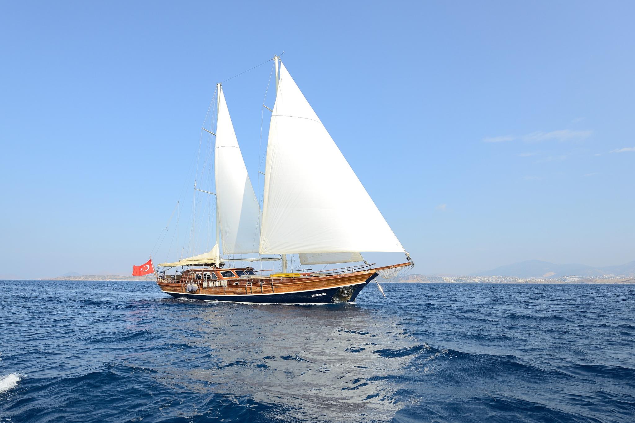 Traditional wooden gulet sailing with white sails raised against clear blue sky and coastline