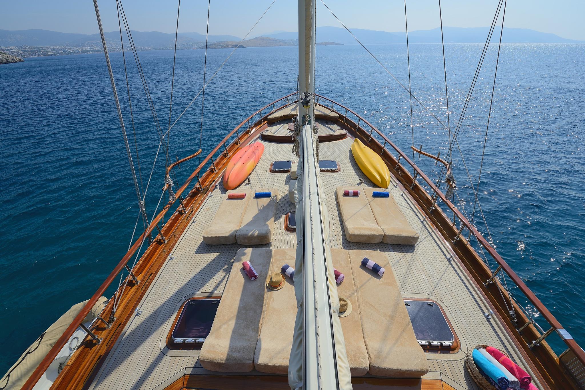 Gulet bow deck showing sun loungers, kayaks, and deck cushions with Mediterranean coastline