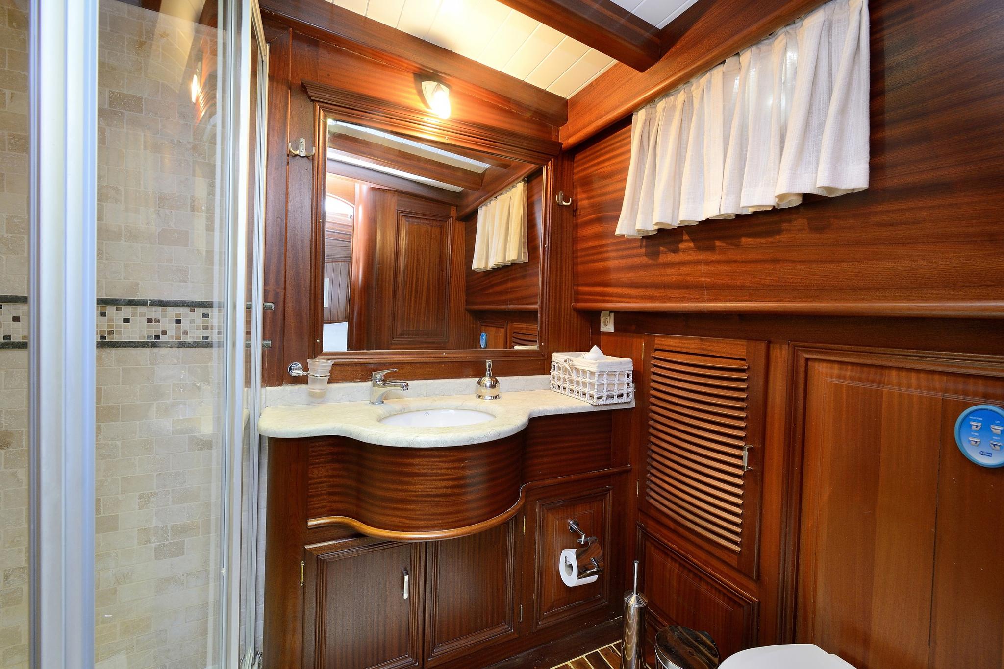 yacht bathroom with rich wood paneling, white marble sink, glass shower enclosure and curtained porthole