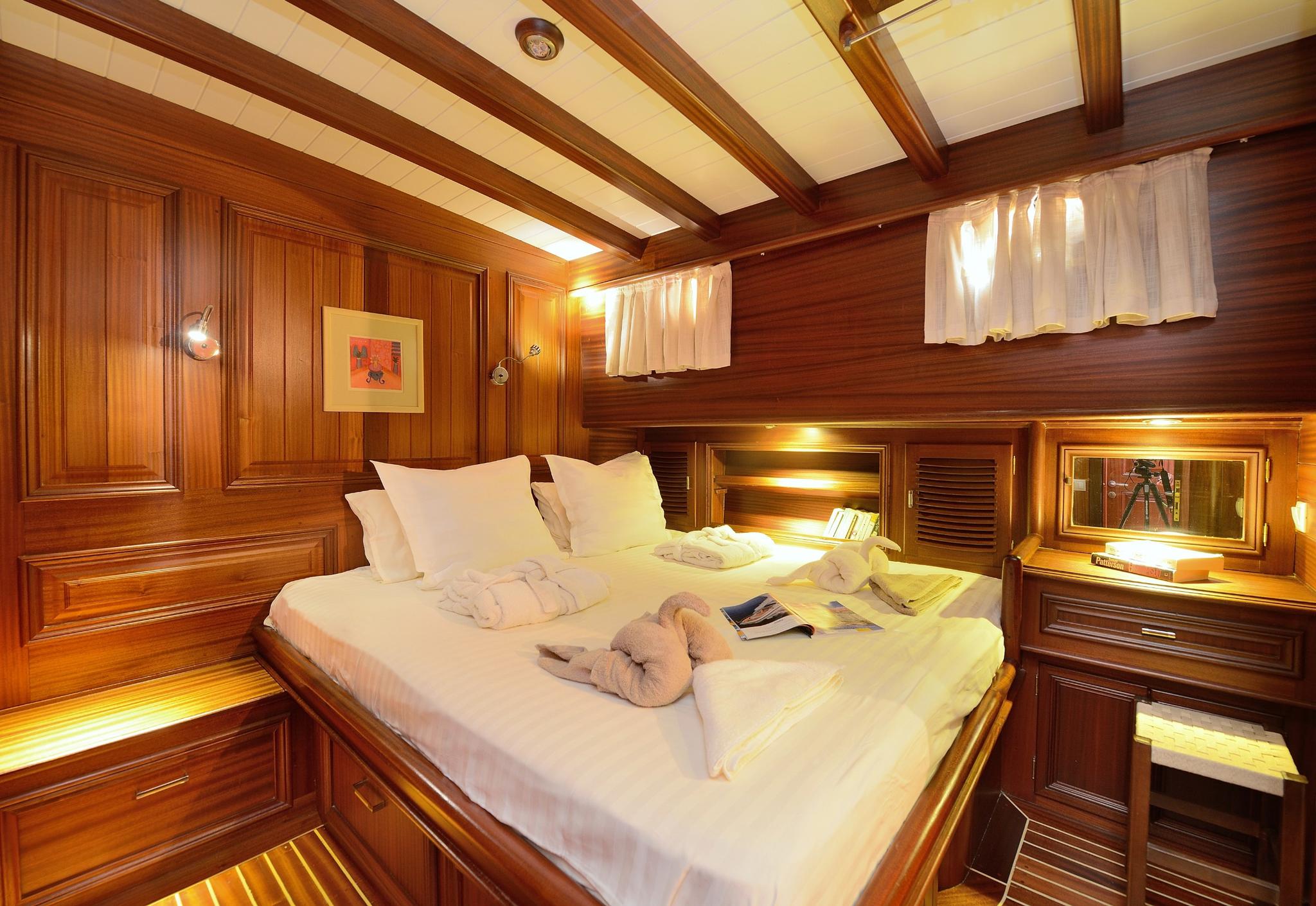 yacht cabin interior with double bed, rich wood paneling, exposed ceiling beams and portholes