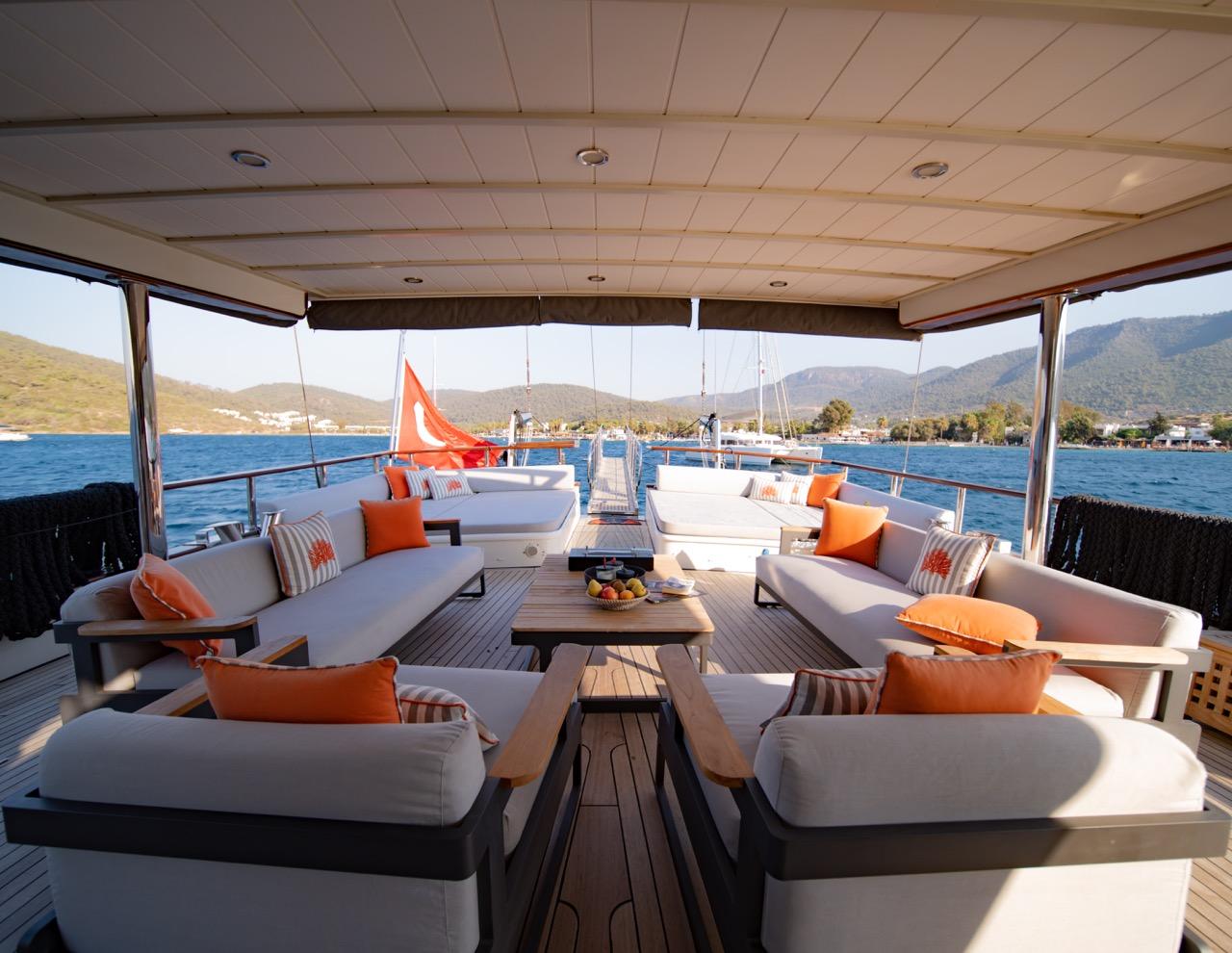 Yacht aft deck with white cushioned seating, orange accent pillows, teak decking and mountain coastline views