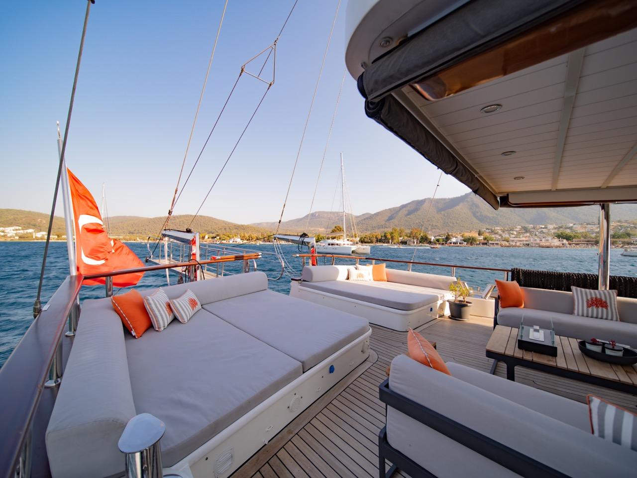 Gulet aft deck with white cushioned seating, dining table, and teak decking overlooking coastal waters