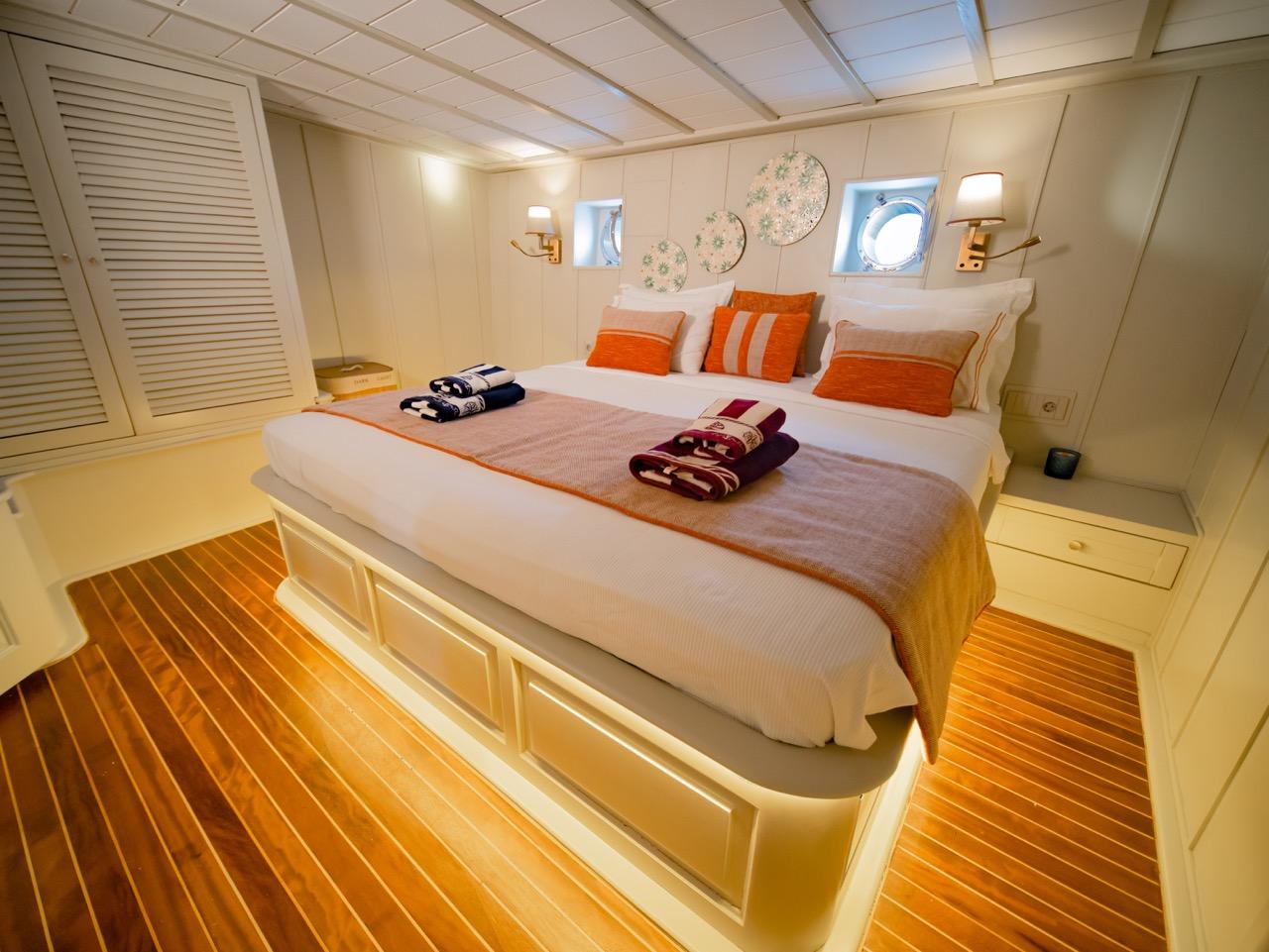 yacht guest cabin featuring double bed with white linens, orange decorative pillows, teak flooring and porthole windows