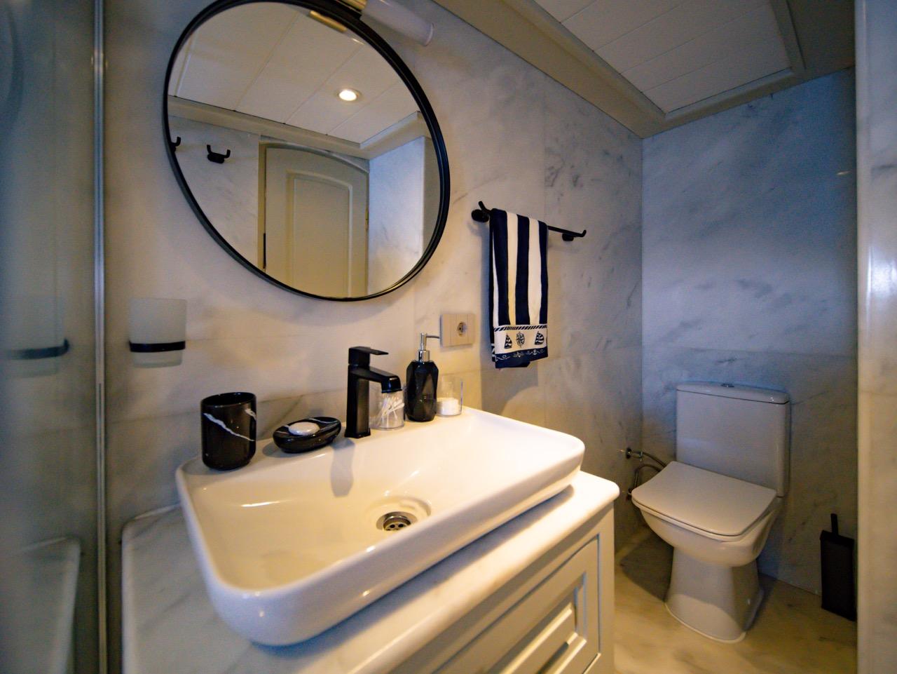 yacht guest bathroom featuring white marble walls, vessel sink, round black mirror and toilet