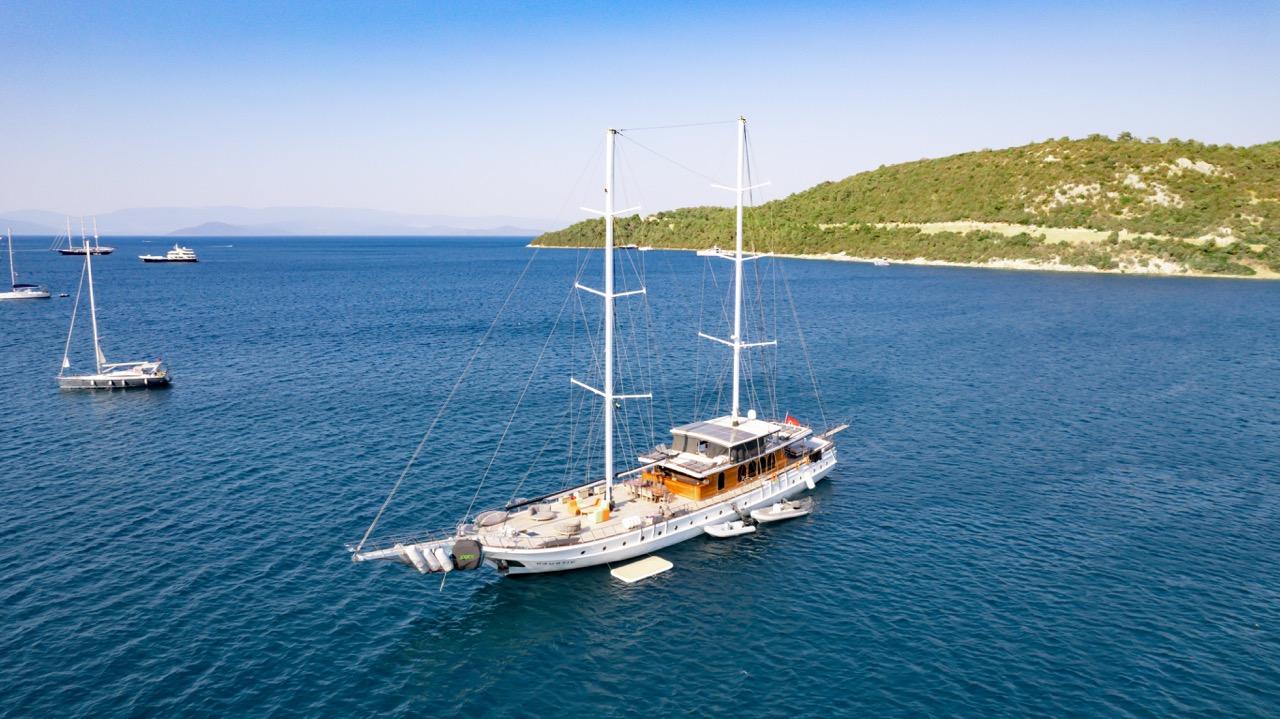 aerial view of white luxury gulet with two masts anchored in turquoise Mediterranean waters near forested coastline