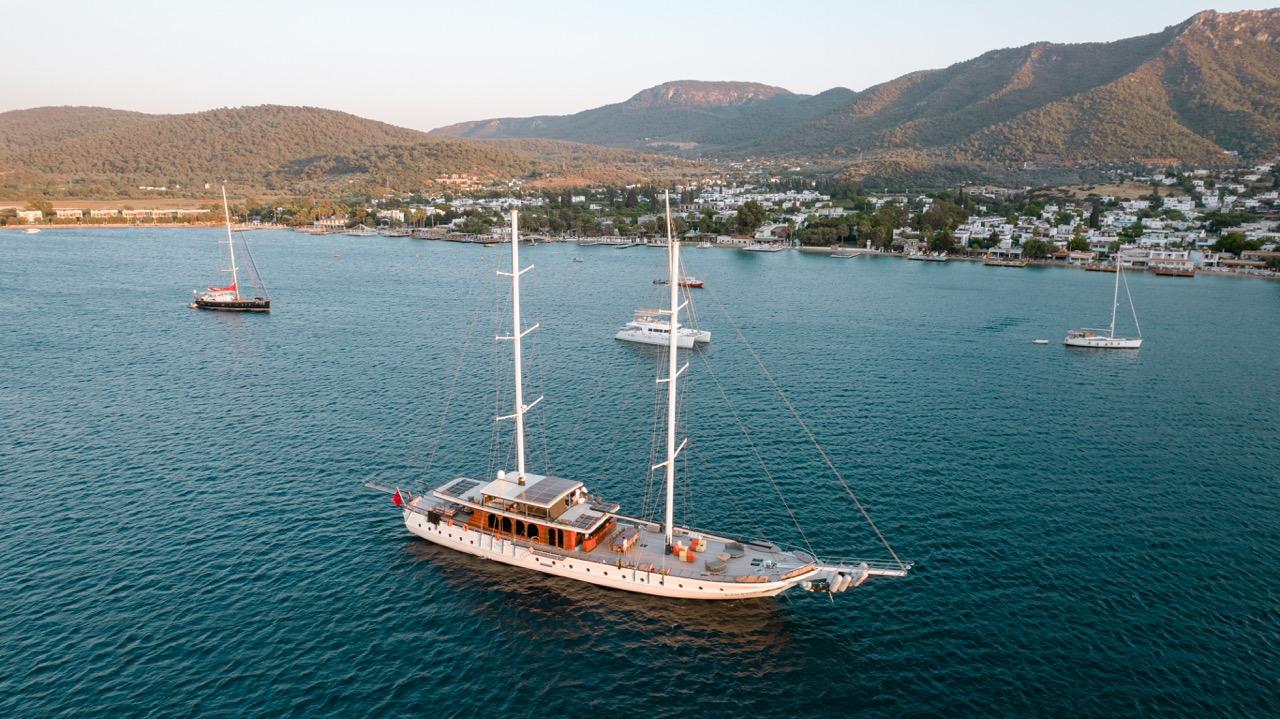 aerial view of white traditional gulet with two masts anchored in turquoise Mediterranean bay surrounded by hills