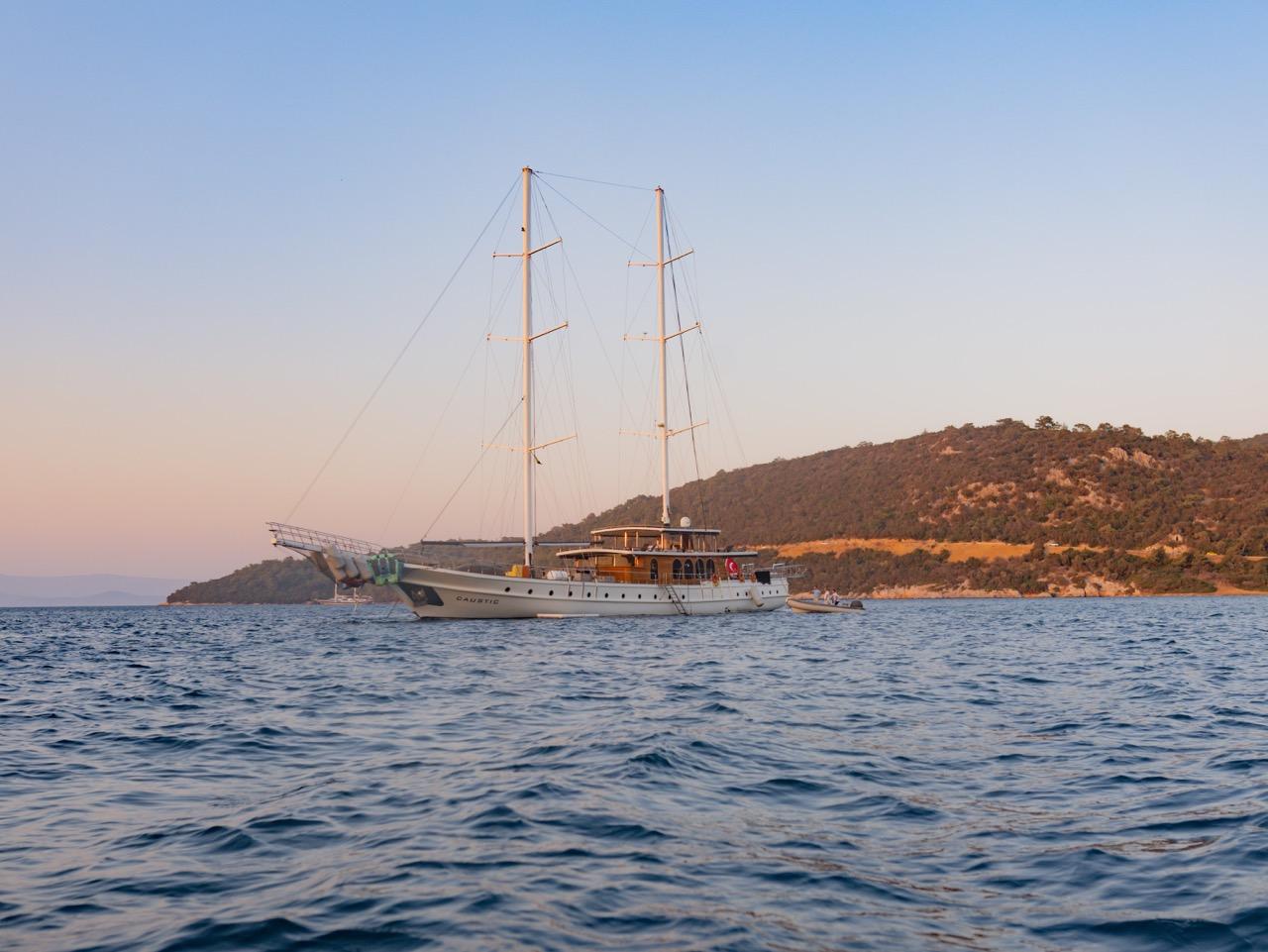 White gulet with two masts and rigging anchored in blue waters near wooded coastline at sunset