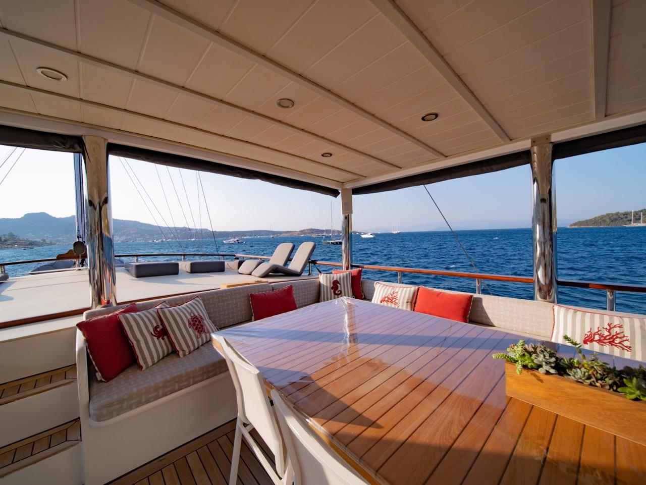 yacht aft deck dining area with built-in seating, teak table, and red cushions overlooking blue sea