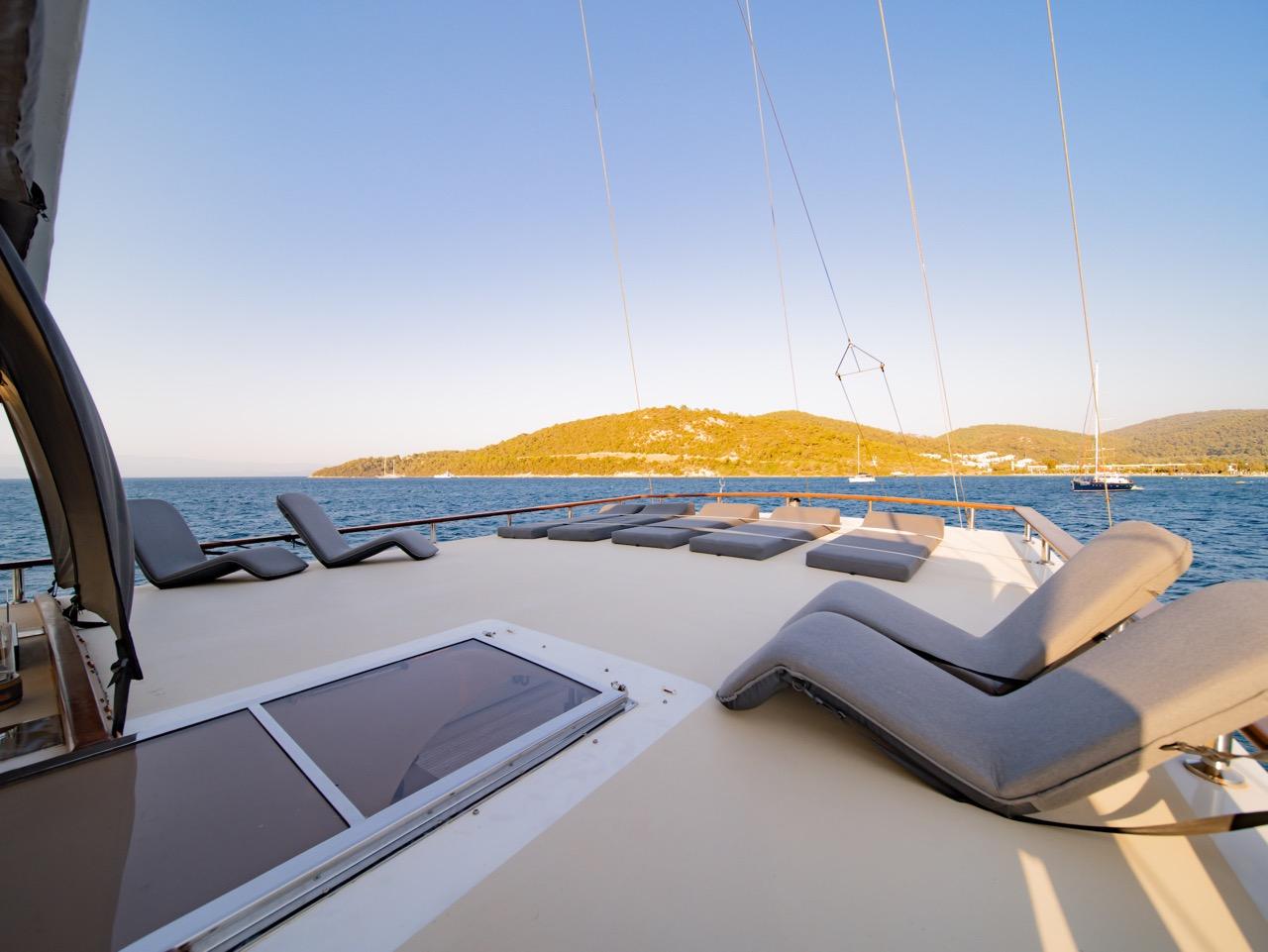 bow sun deck with grey cushioned loungers and yacht rigging against coastal backdrop