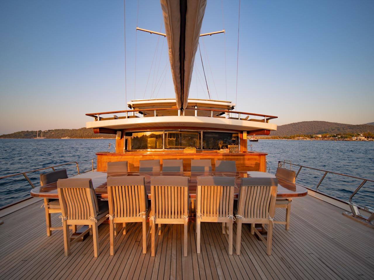 gulet aft deck dining area with wooden table, chairs and wheelhouse at sunset