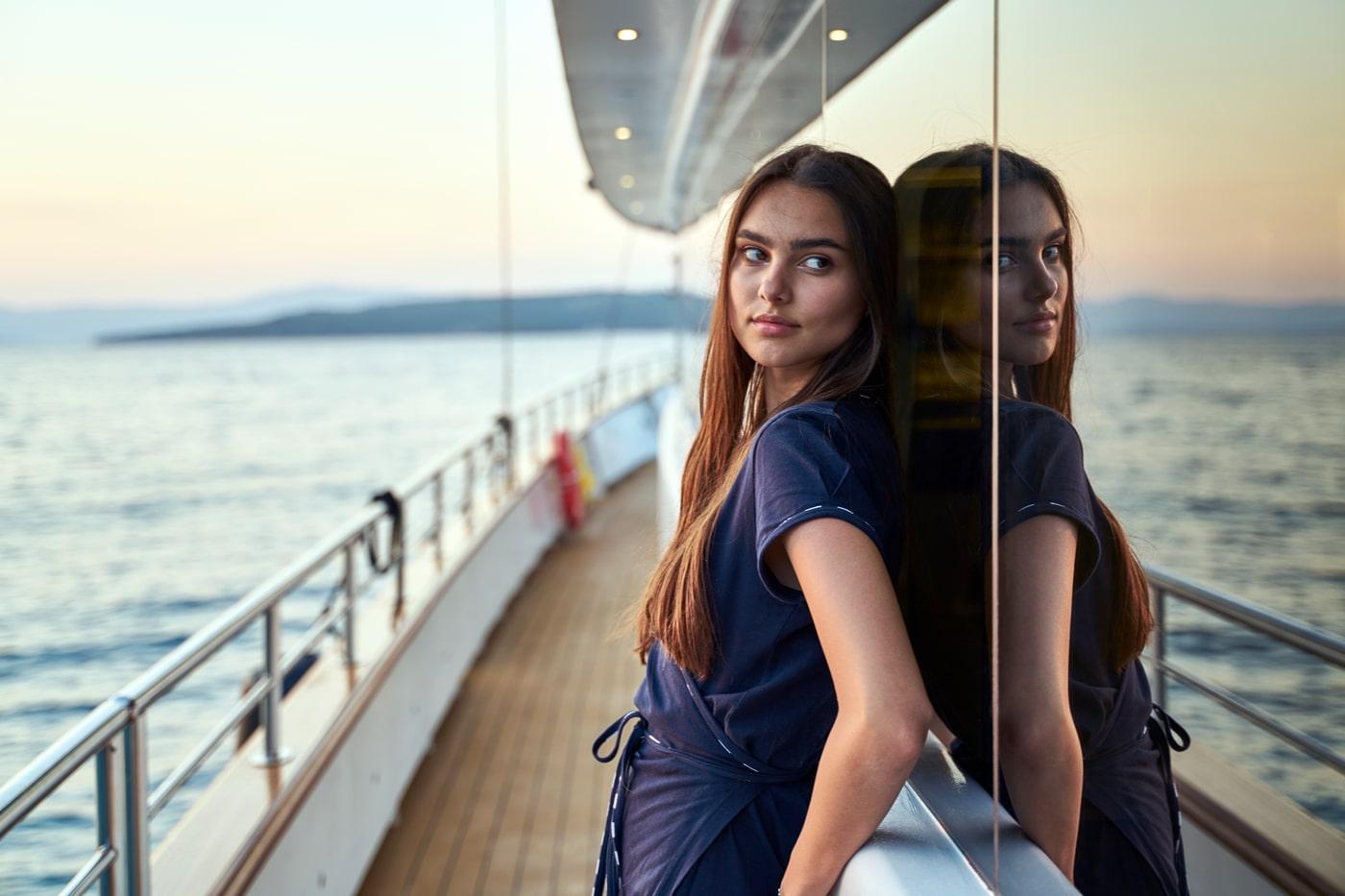 Woman leaning against yacht's glass exterior on teak deck during golden hour