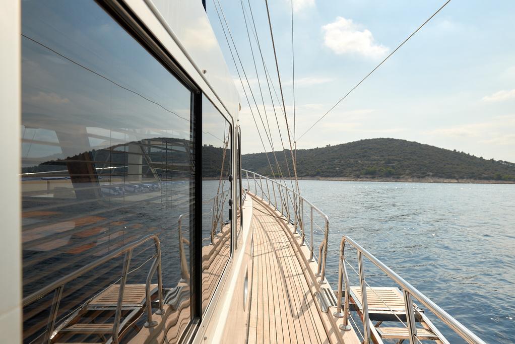 Luxury yacht port side teak deck walkway with glass salon windows and coastal backdrop
