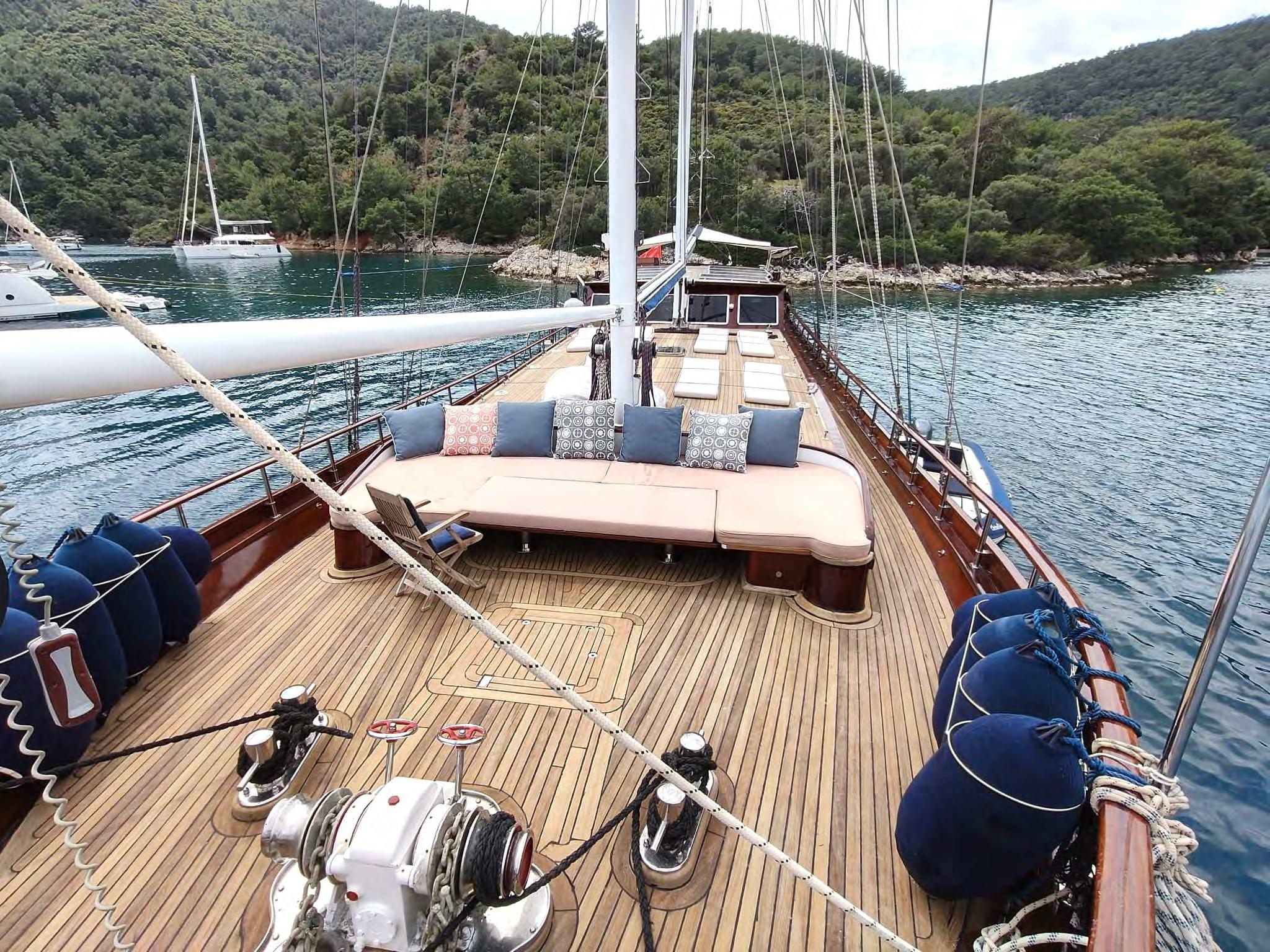 Gulet bow deck featuring large cushioned seating area with blue and patterned throw pillows on polished teak deck, moored in a forested cove