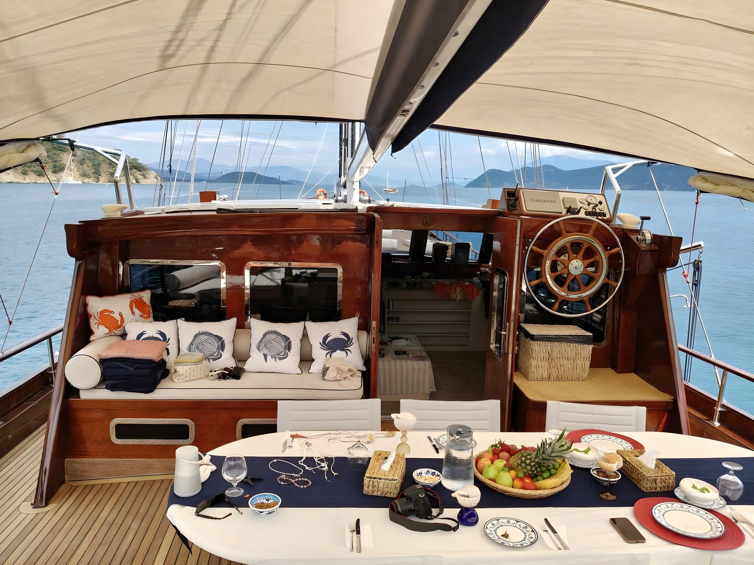 Gulet aft cockpit dining table laid with fresh fruit bowl, glassware, and blue table runner, with cushioned seating and wooden helm station behind