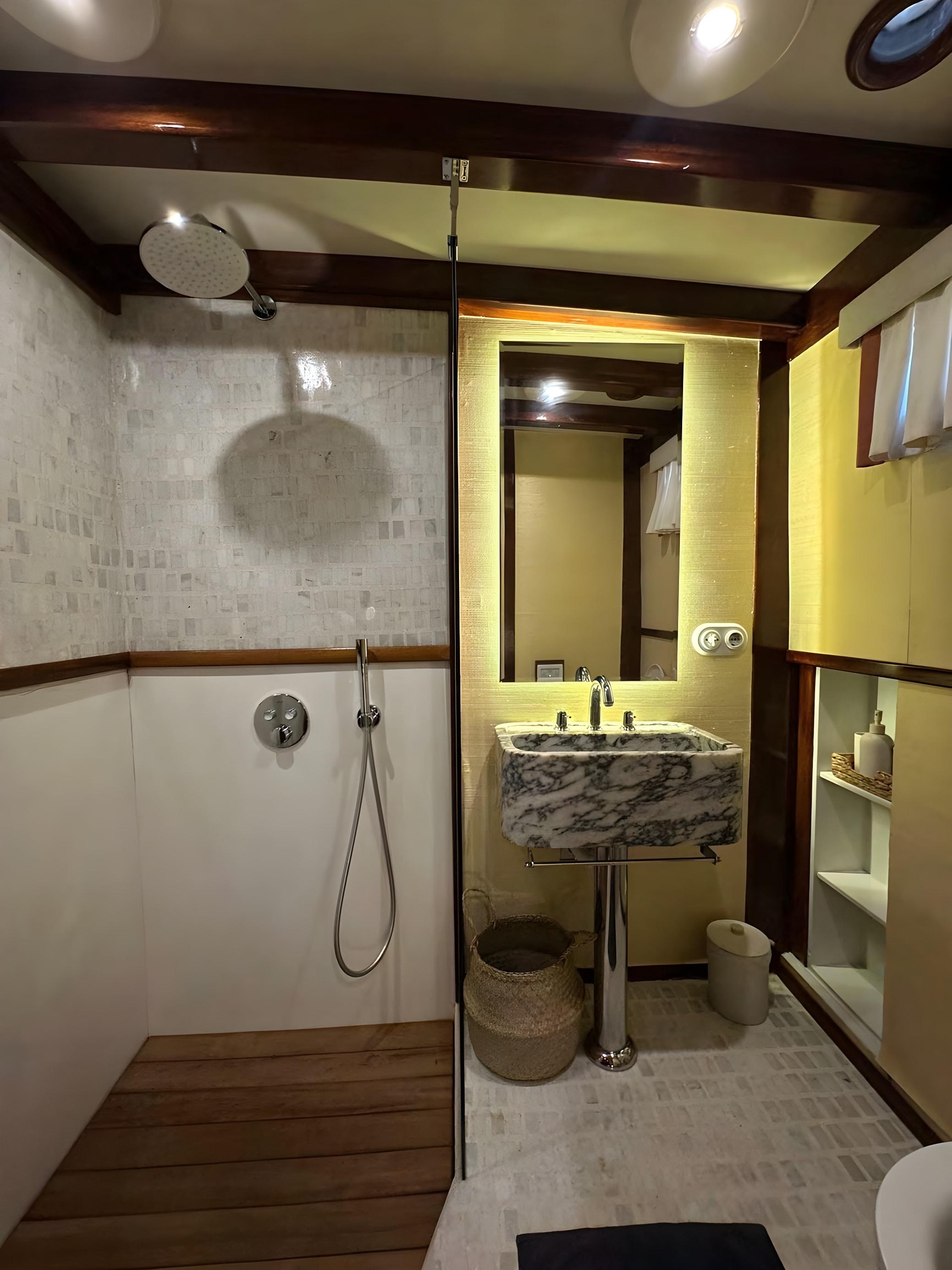 Gulet en-suite bathroom featuring large rain shower head, teak shower floor, marble pedestal sink with backlit mirror, and mosaic tile walls
