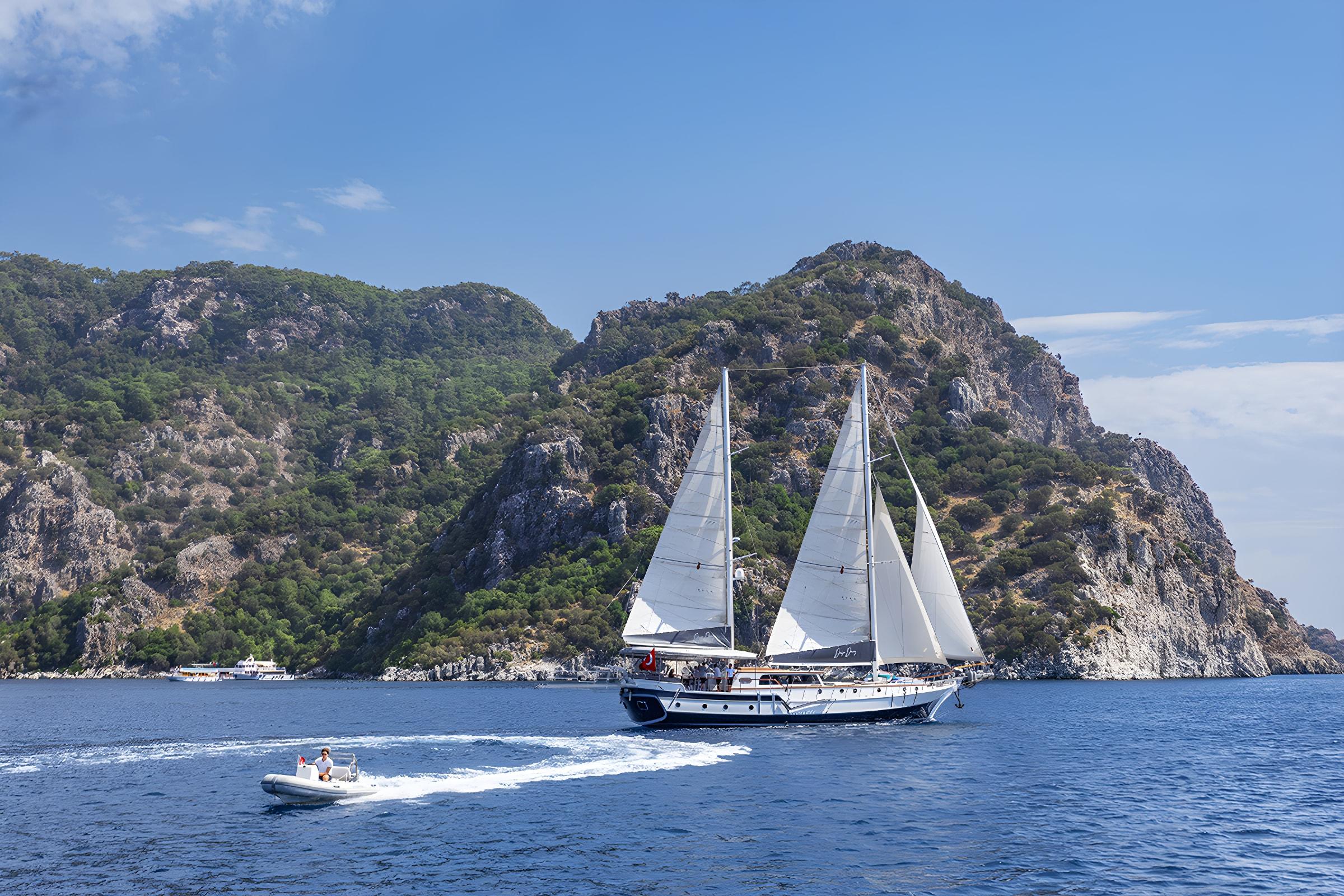 luxury gulet sailing under full white sails with tender RIB alongside rocky Mediterranean cliffs