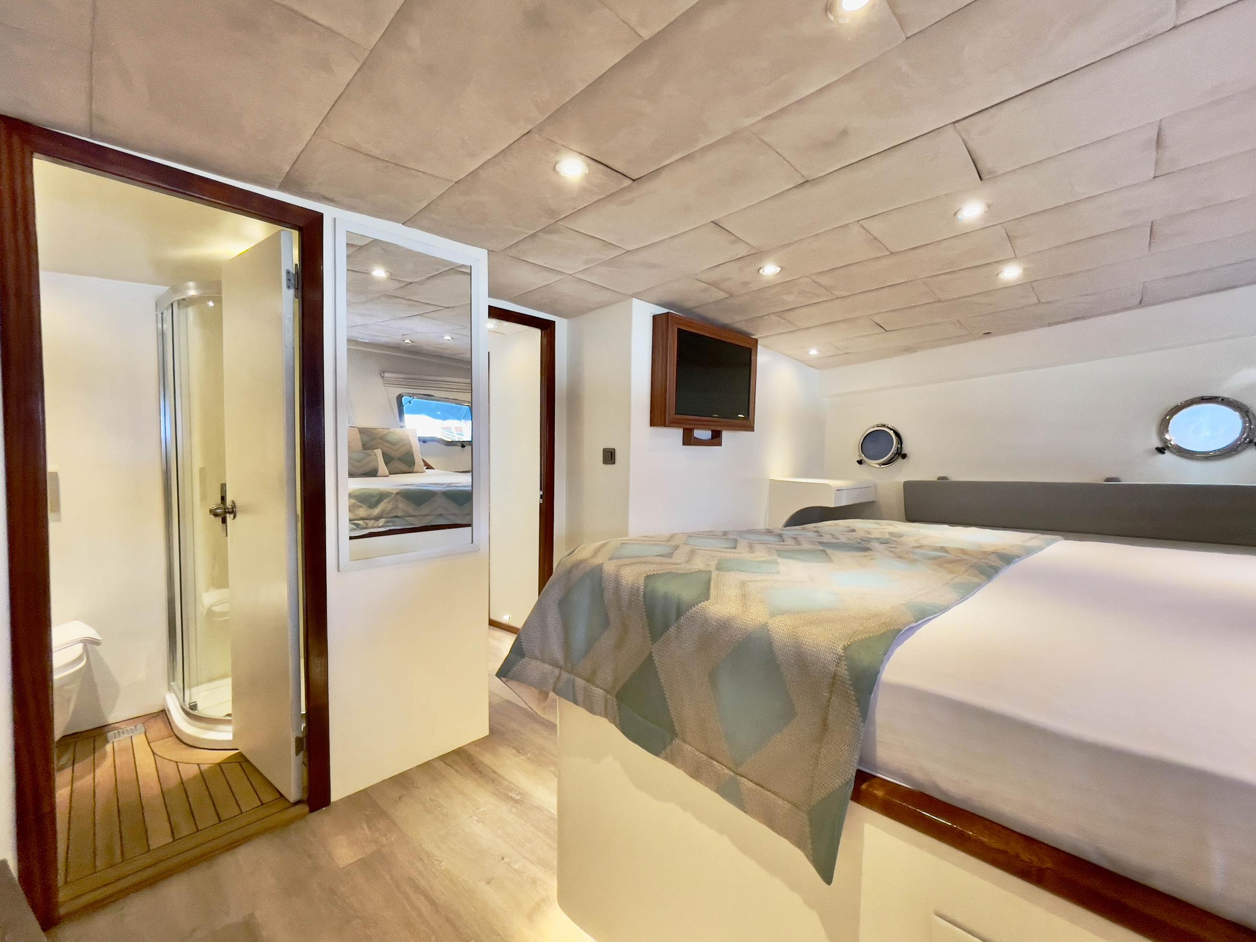 yacht double cabin with quilted bedcover, wall-mounted flatscreen TV, two portholes, and open en-suite shower and WC