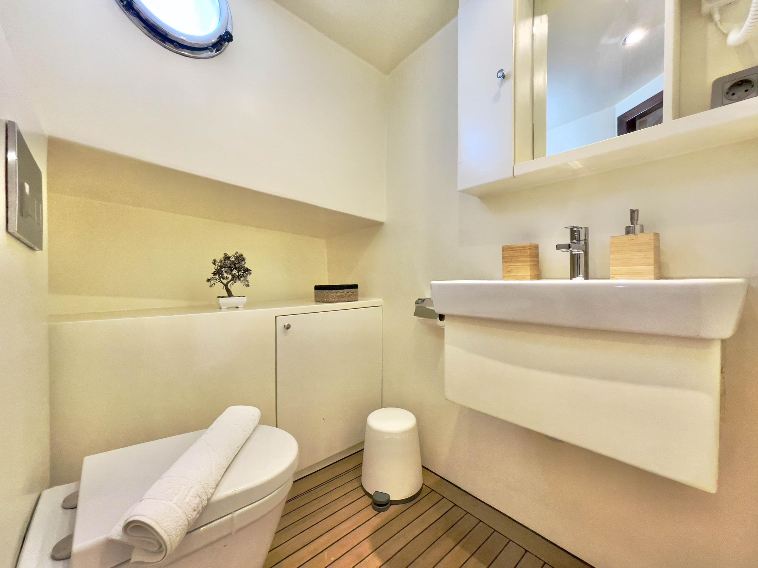 Yacht en-suite bathroom with wall-mounted basin, chrome tap, teak sole floor, toilet with folded white towels, storage cabinet, and circular porthole
