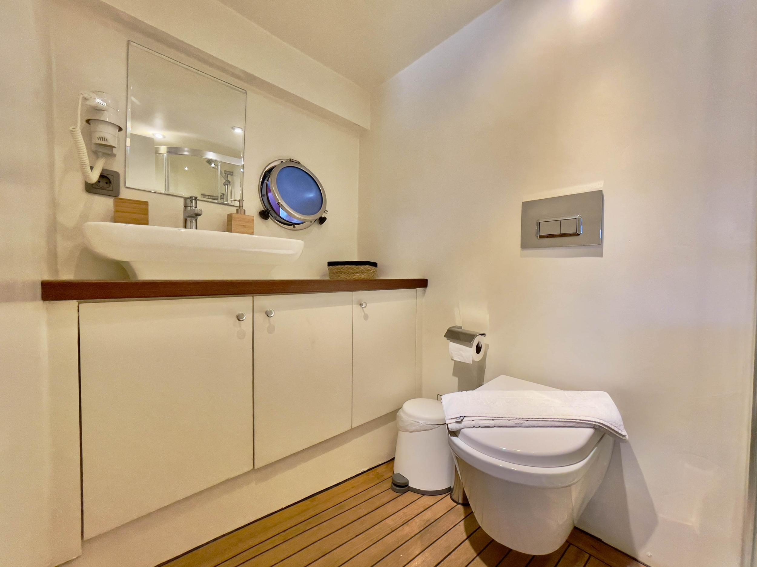 Yacht en-suite bathroom with vessel porthole, white vanity unit, teak floor, and wall-hung toilet