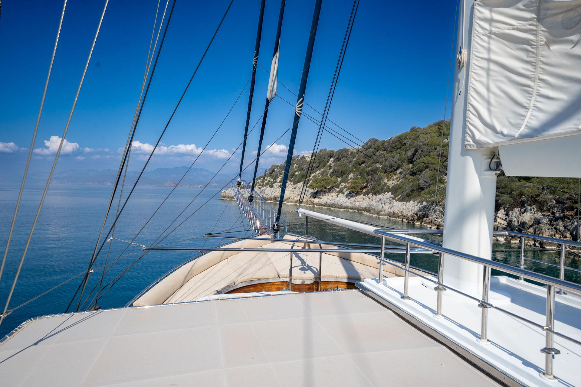 gulet forward deck with white railings, mast rigging, and Mediterranean coastline view