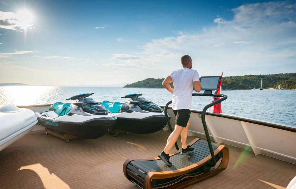 Guest using curved treadmill on luxury yacht deck with jet skis nearby
