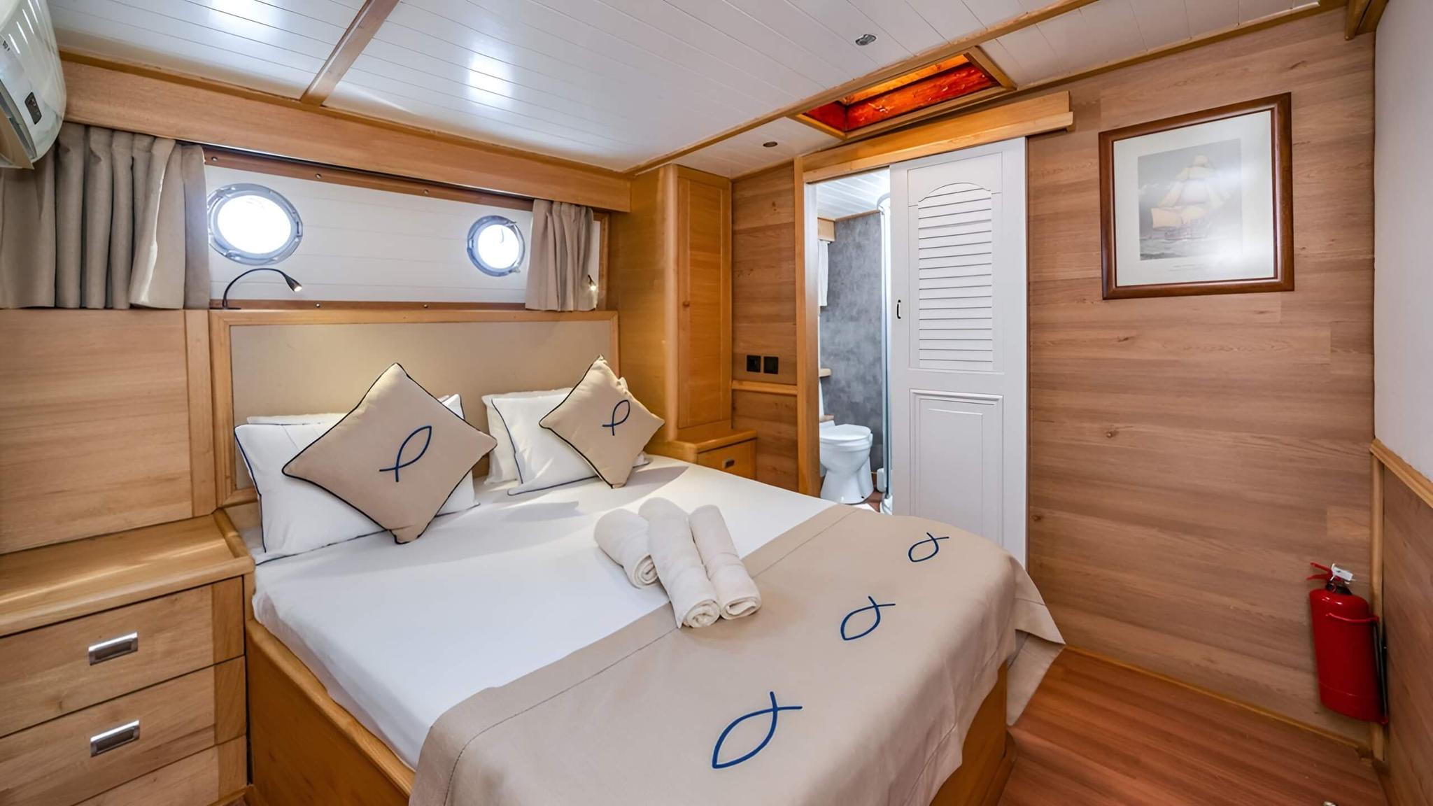 yacht guest cabin featuring double bed with beige linens, two portholes, built-in storage and en-suite bathroom