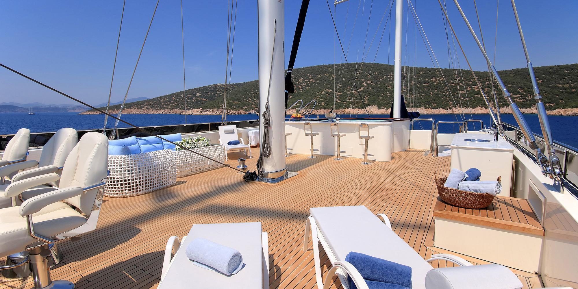 gulet main deck with teak decking, white sun loungers, central mast and Mediterranean coastline