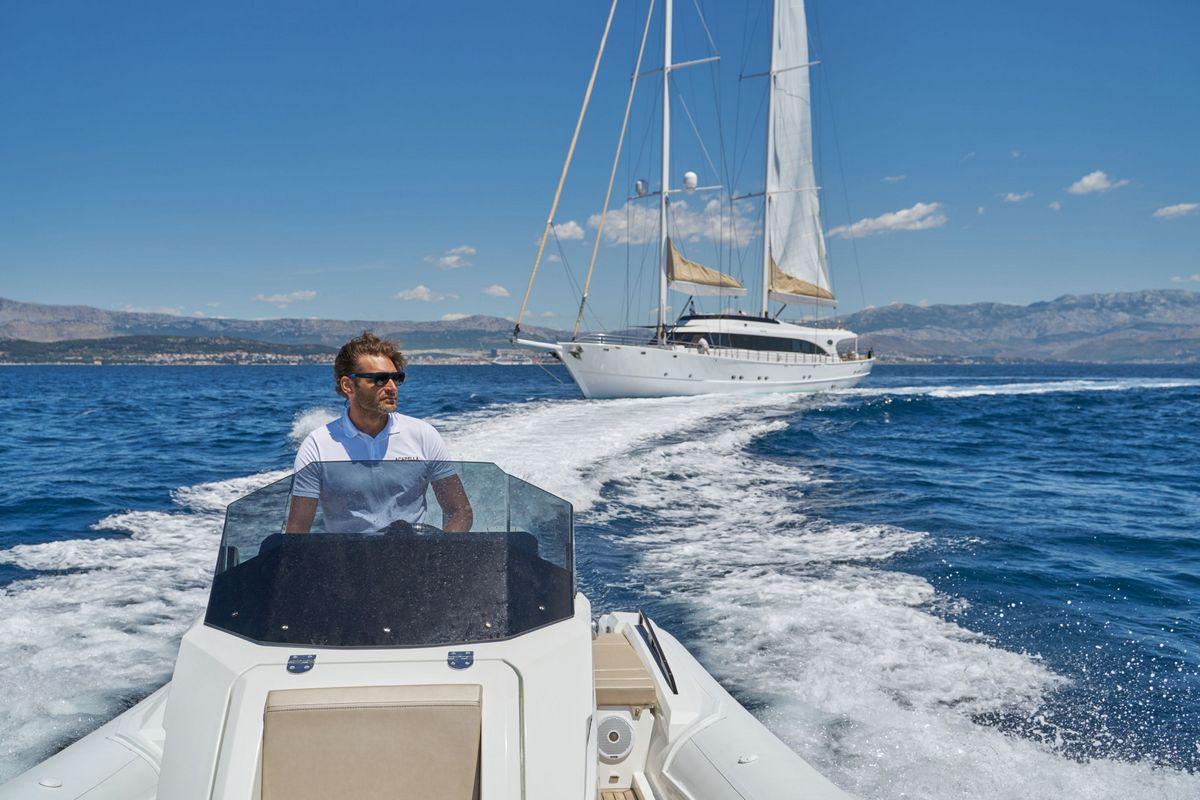 Tender boat operator with white sailing yacht creating wake in blue waters
